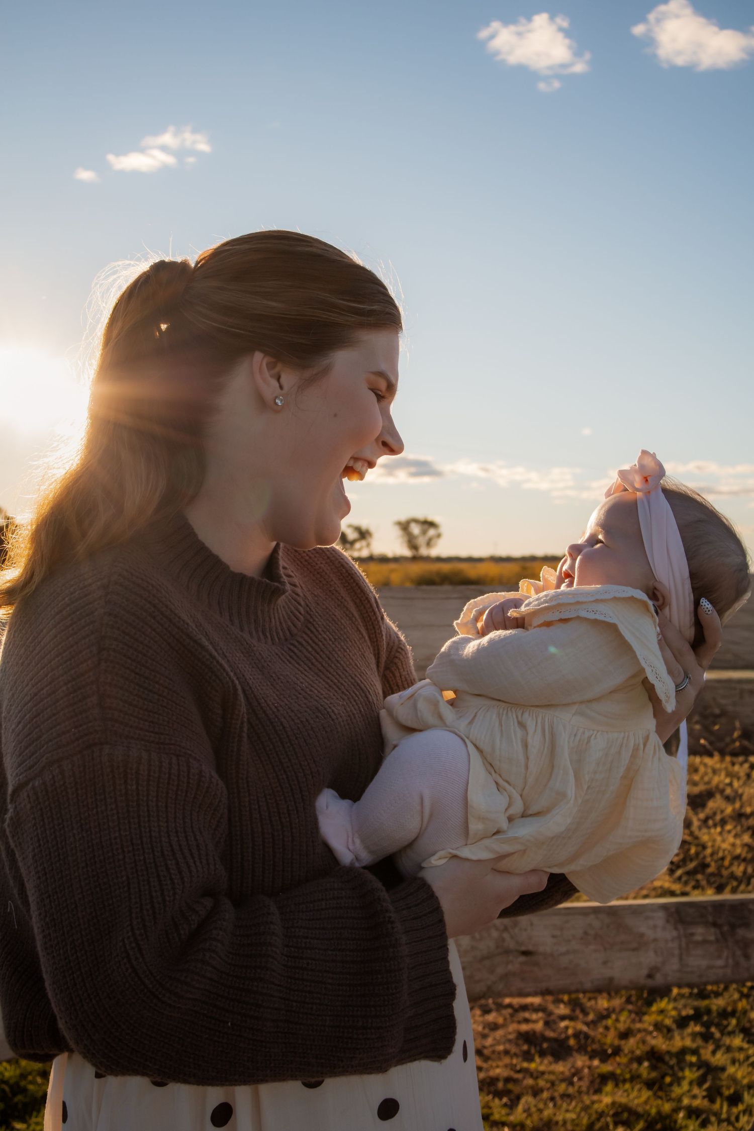 A mother holds her smiling baby outdoors at golden hour, both dressed warmly, with a sunlit rural landscape behind them.