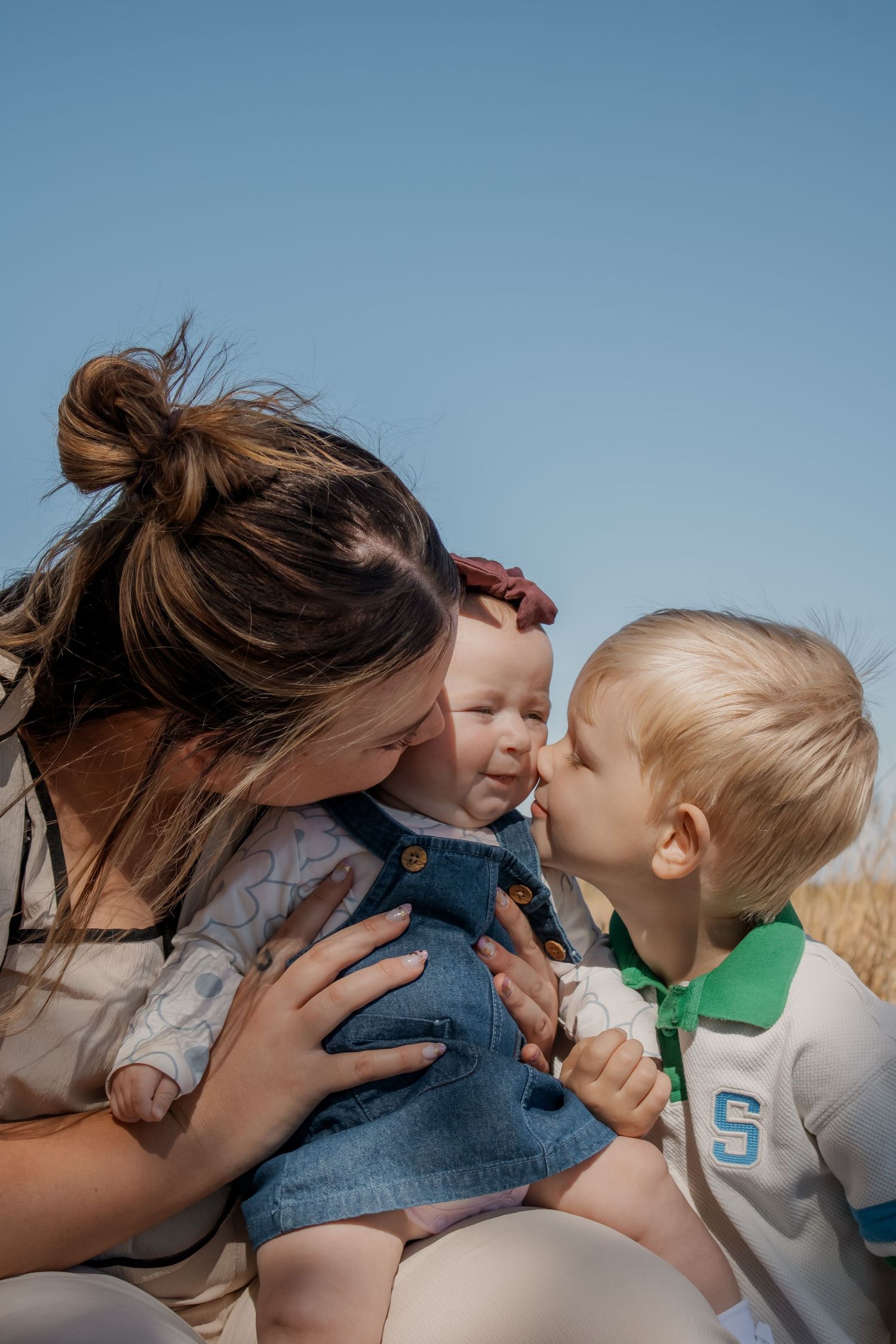 A mother holds her baby girl while a young toddler boy leans in to give the infant a sweet kiss outdoors.