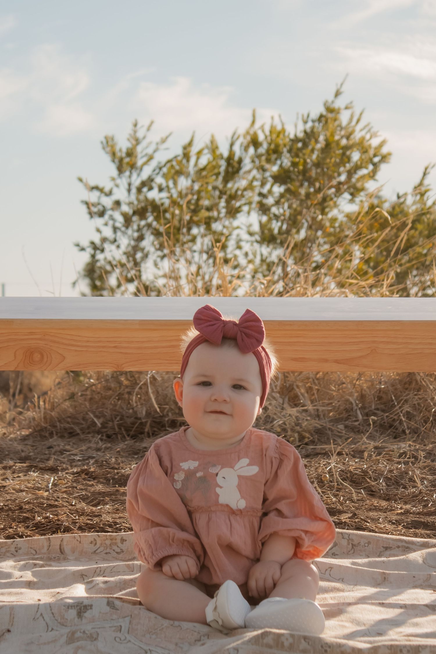 Adorable baby girl in pink bunny romper and bow headband sitting outdoors on a blanket with greenery background.