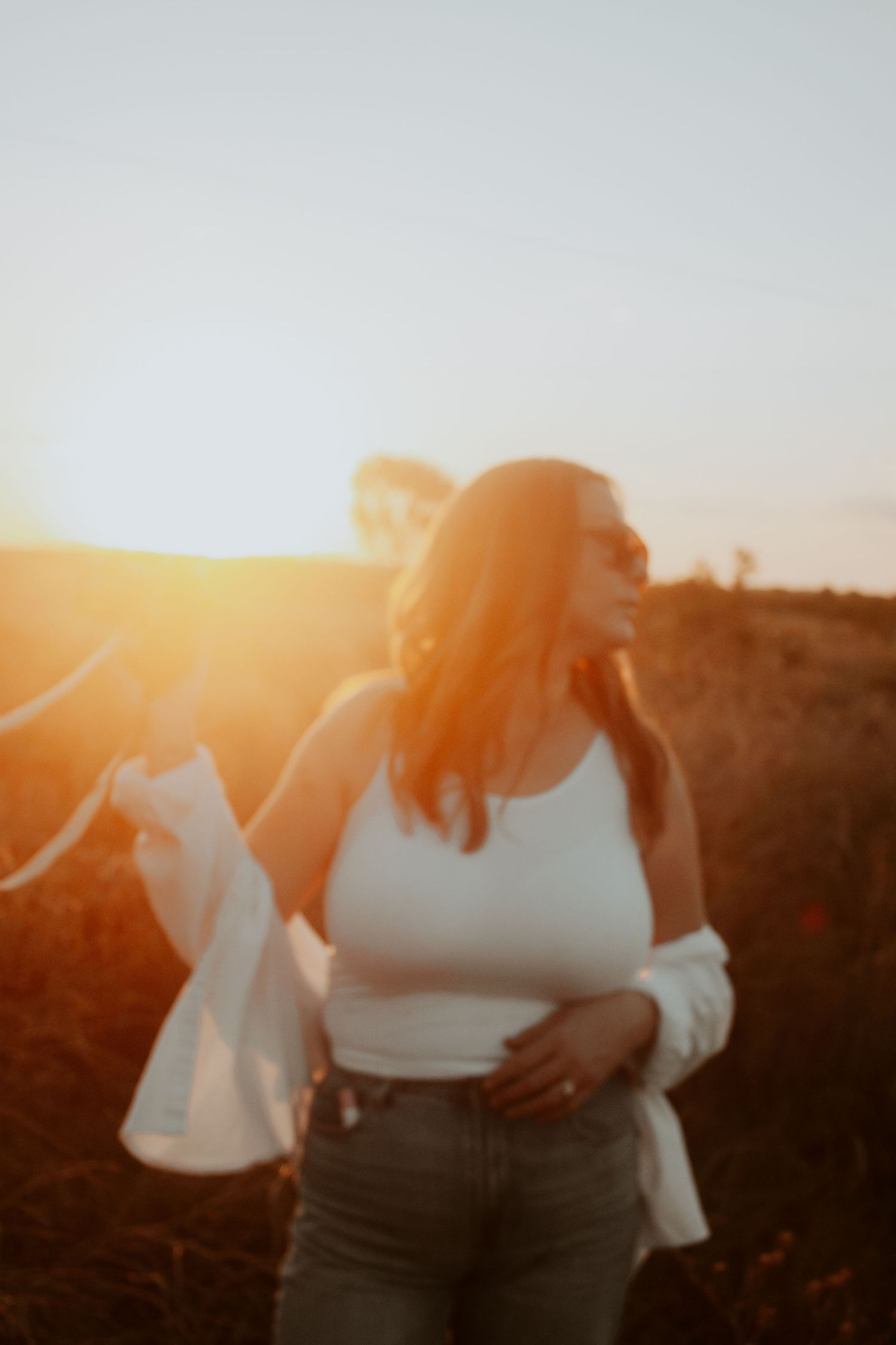 Woman in white tank top smiling at golden sunset in a field, holding open shirt.