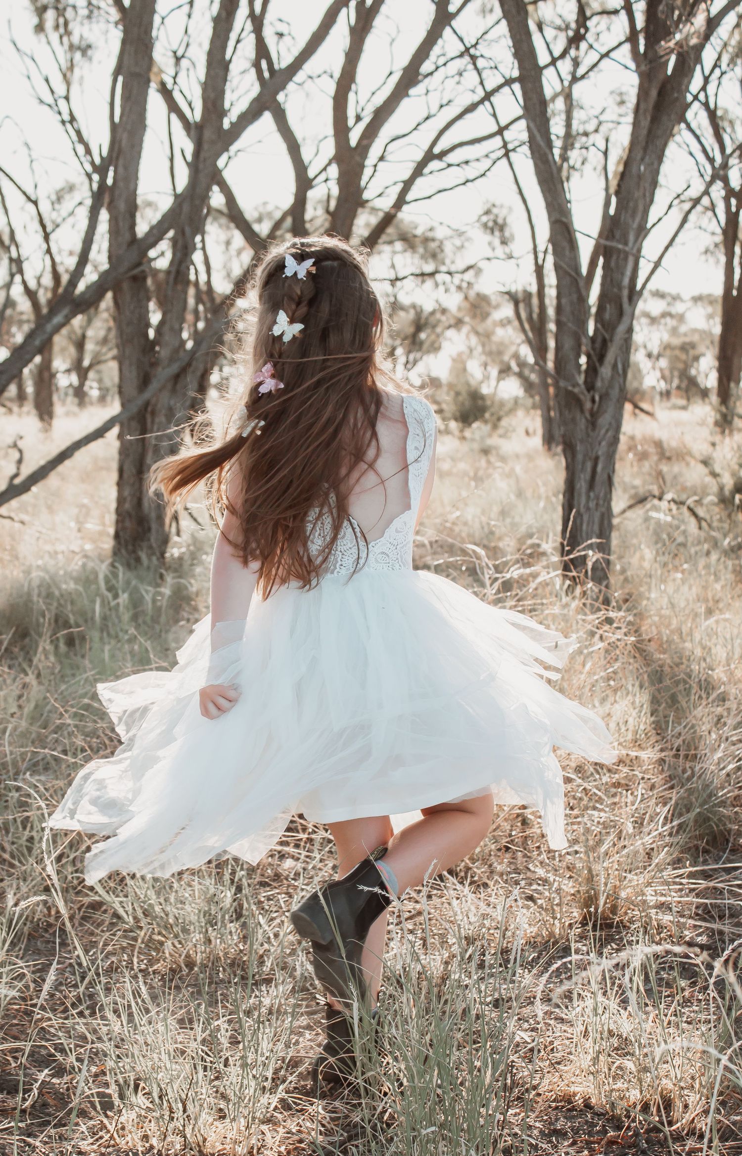 Girl in white tulle dress and cowboy boots twirling among bare trees in a field, butterfly hair accessories.