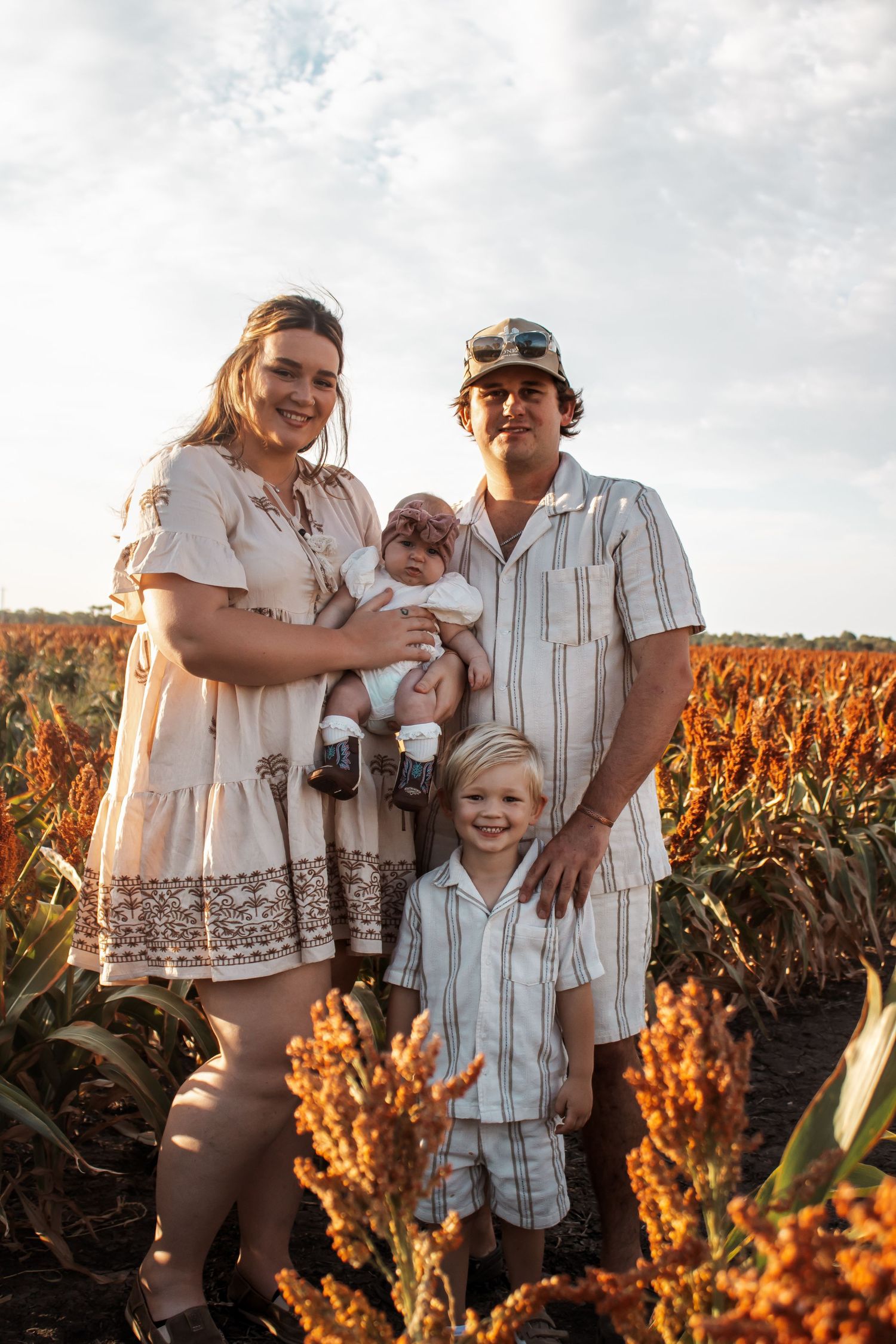 Family of four posing in a golden sorghum field, parents holding a baby with a toddler standing in front.