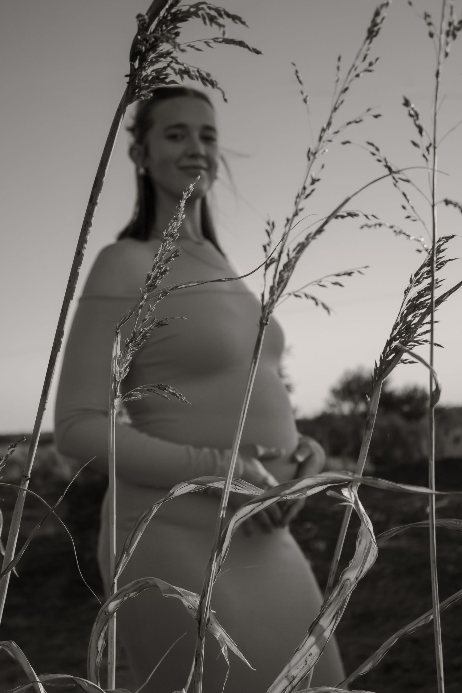 Black and white photo of a pregnant woman standing in tall grass, cradling her baby bump outdoors.