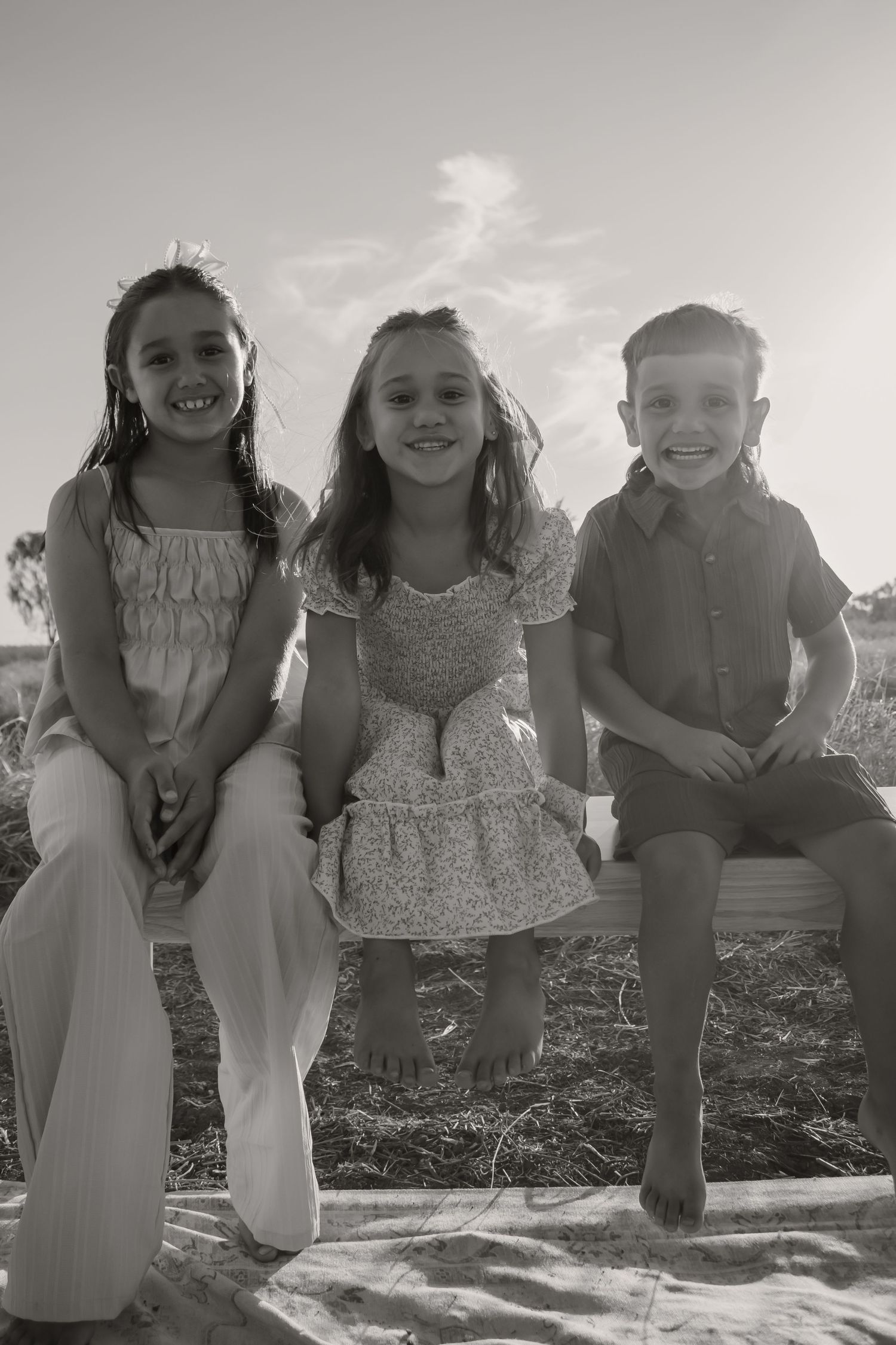 Three smiling children sitting together outdoors in a black and white photo on a sunny day.