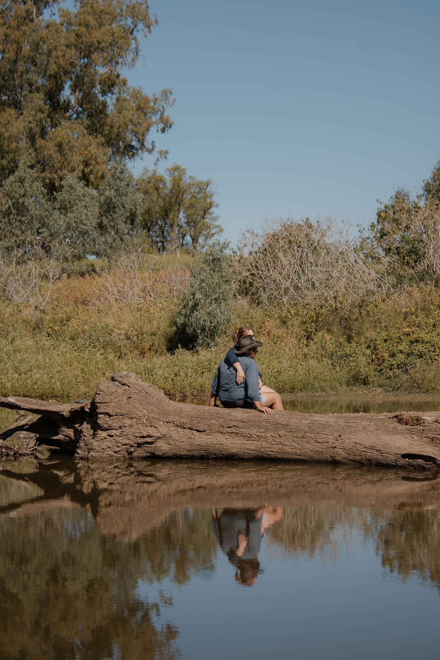 Person sitting on a fallen log beside a calm reflective river surrounded by lush green trees and shrubs on a sunny day.