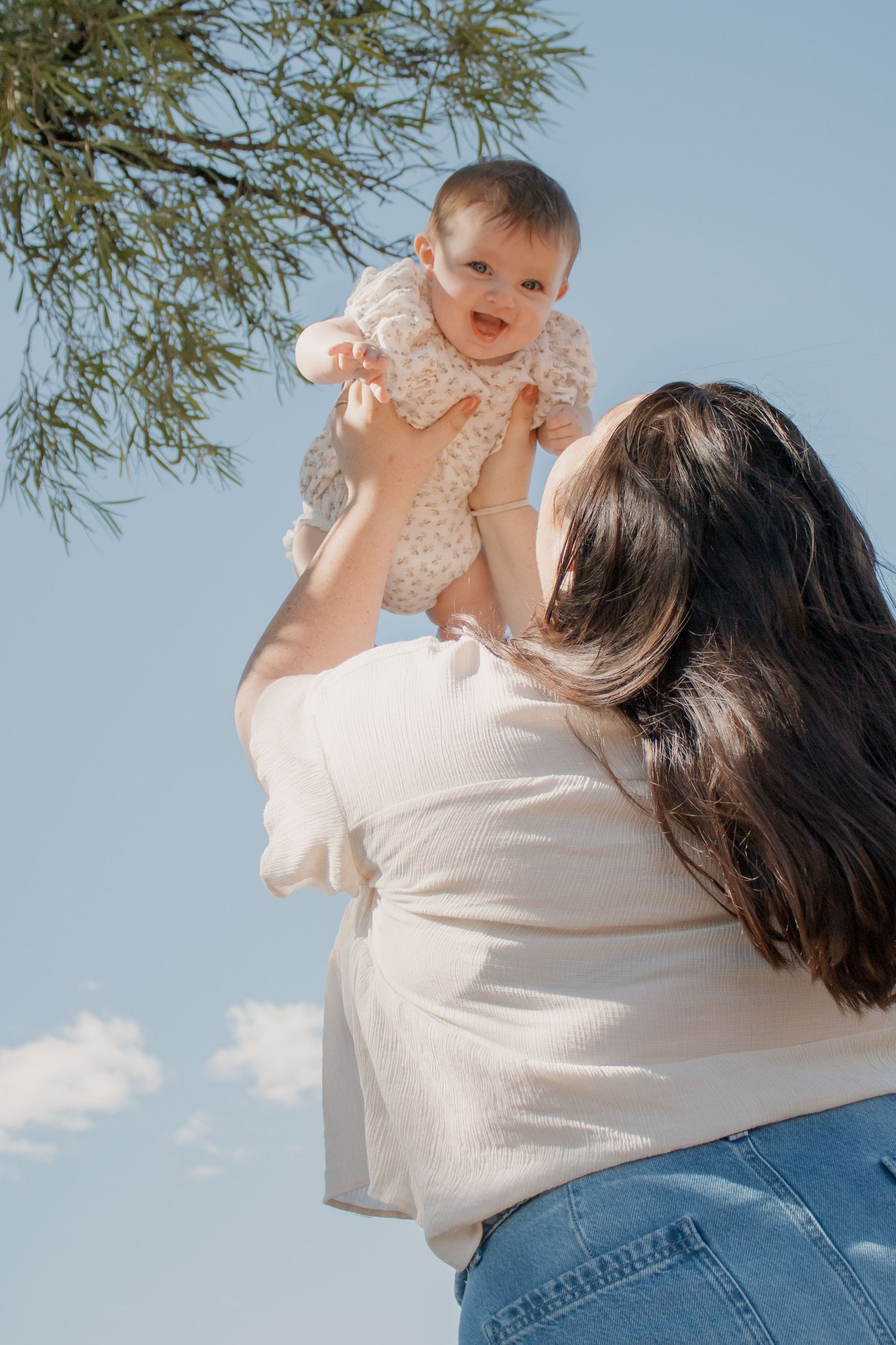 A mother lifts her smiling baby girl into the blue sky outdoors, sharing a joyful moment beneath a green tree.