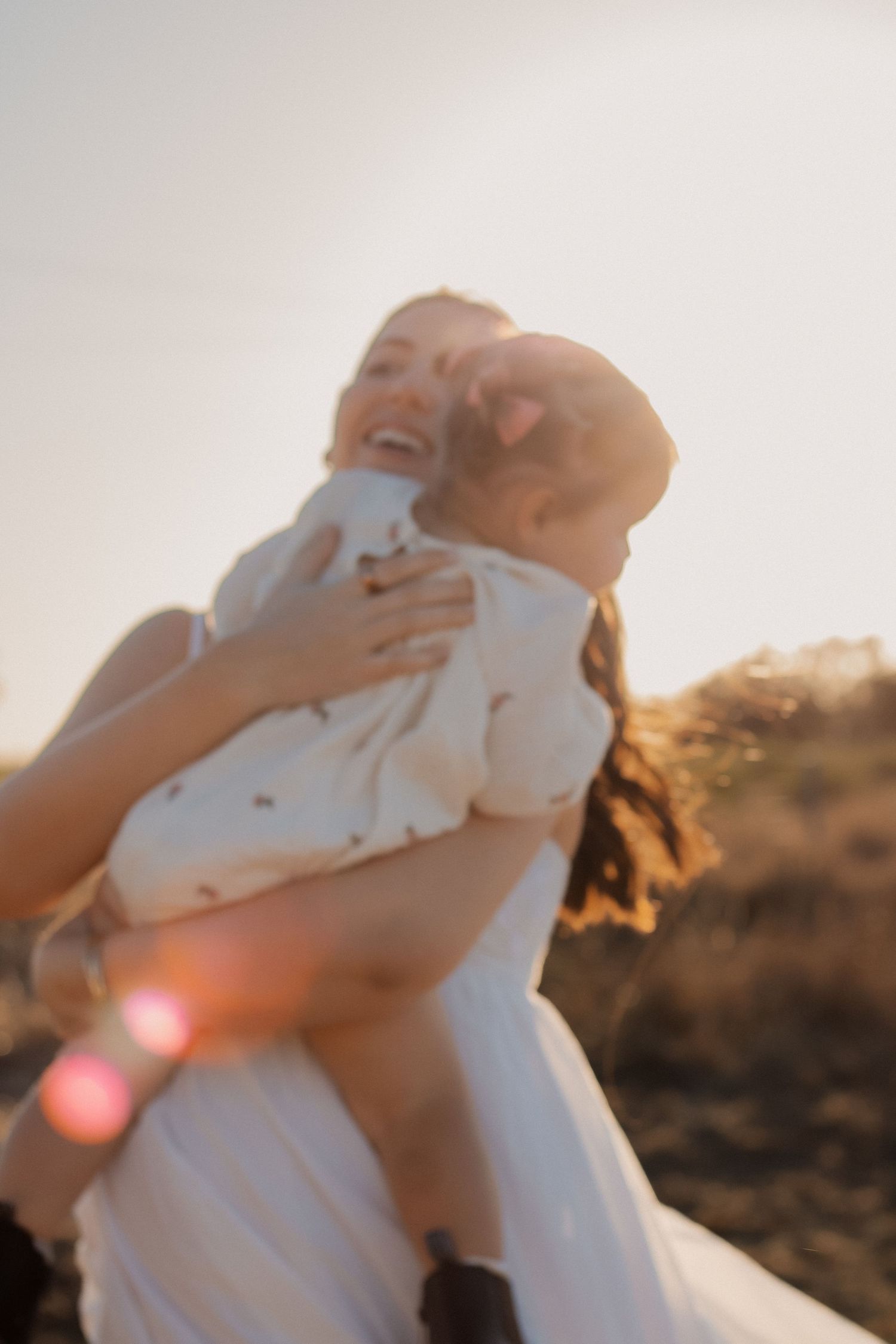 A joyful mother laughs while holding her baby outdoors during golden hour sunset light.