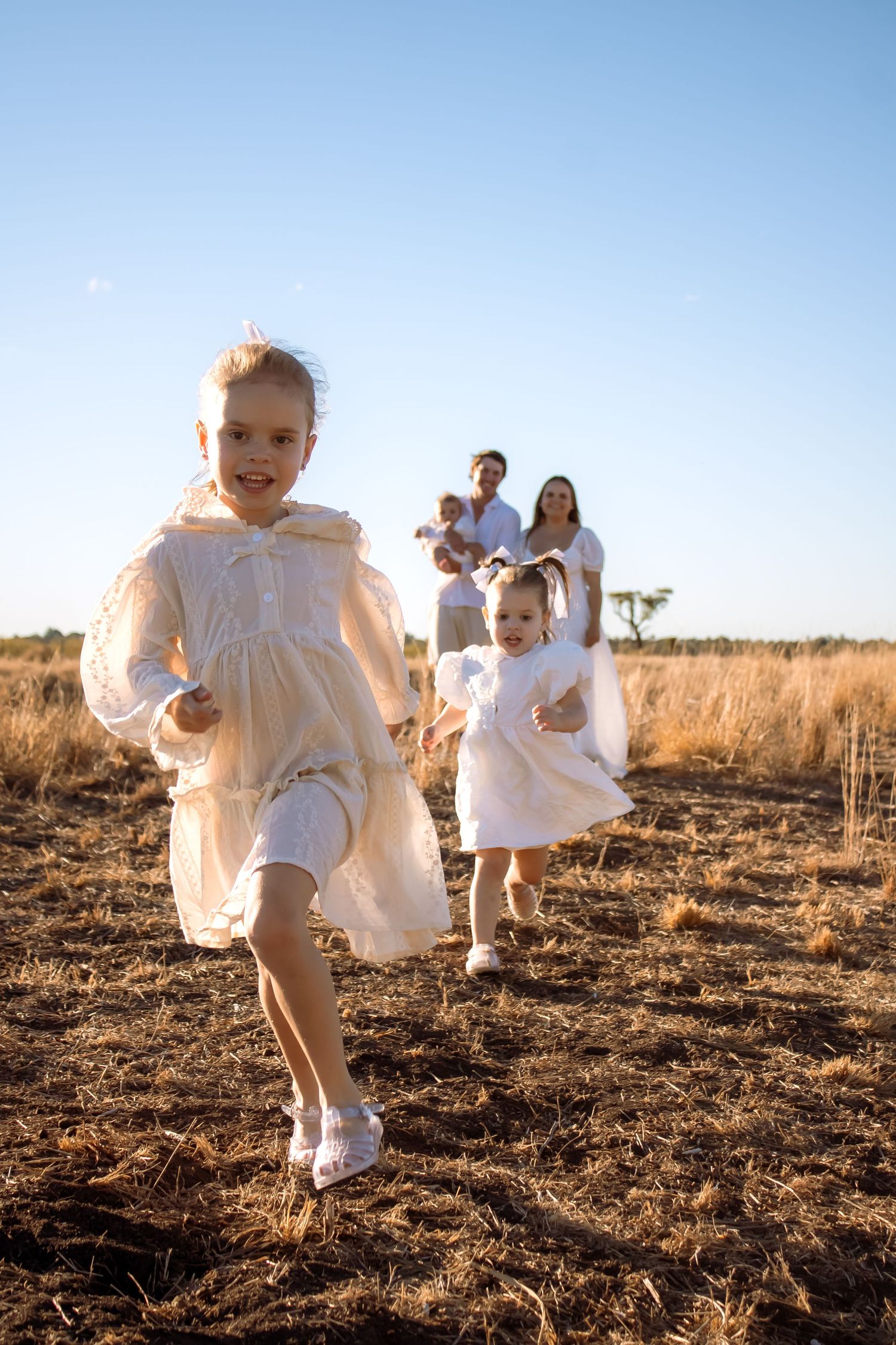 Two young girls in white dresses run through a golden field while their parents stand smiling in the background.