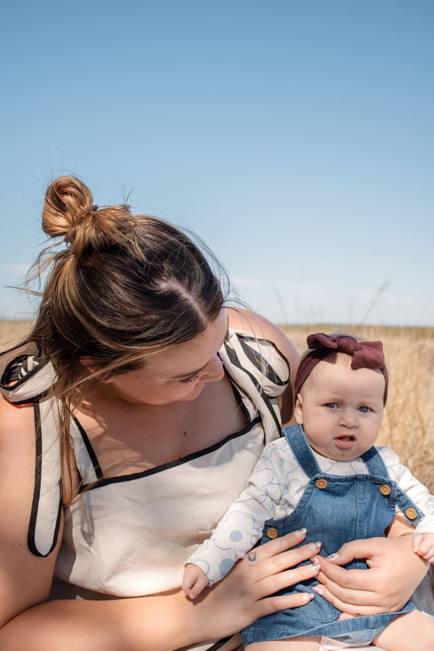 A mother tenderly holds her baby dressed in denim overalls outdoors in a golden field under a clear blue sky.