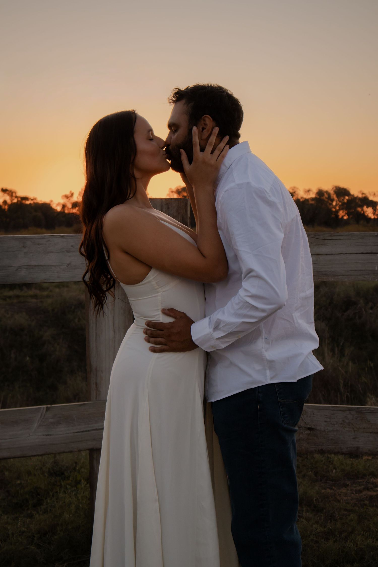 Couple sharing a romantic kiss at sunset near a rustic wooden fence in a golden field.
