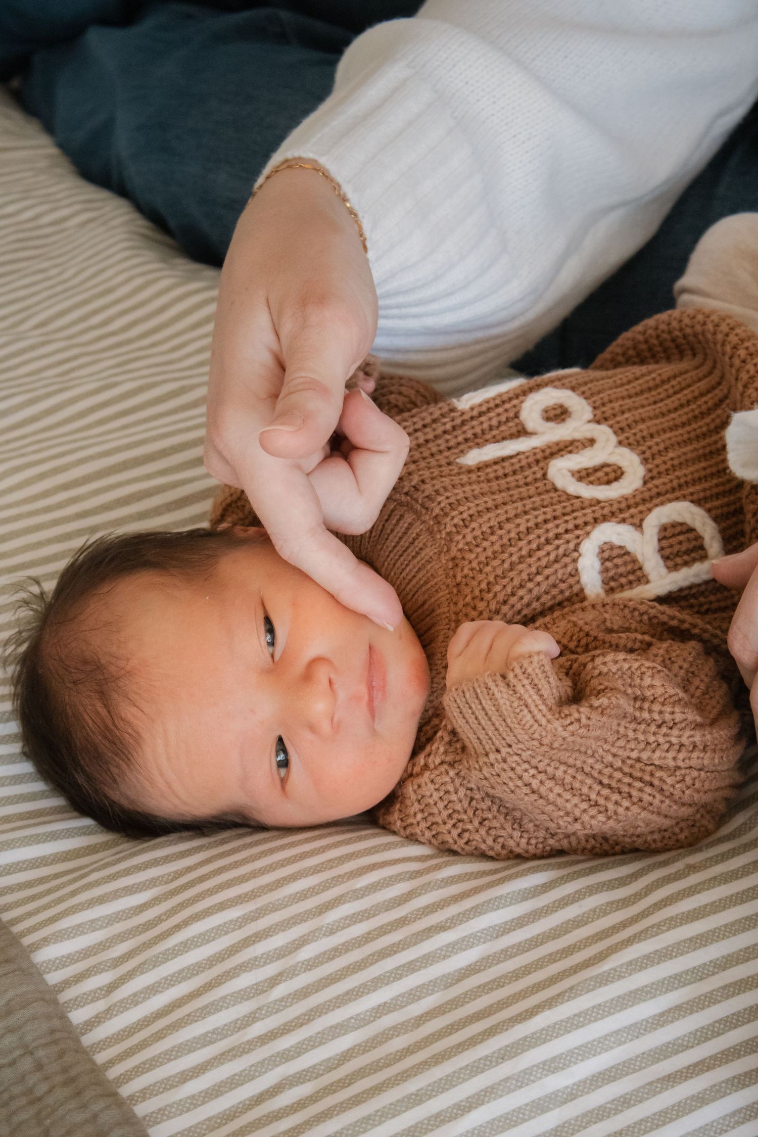 Newborn baby in brown knit sweater lying on striped blanket while parent gently touches their head.