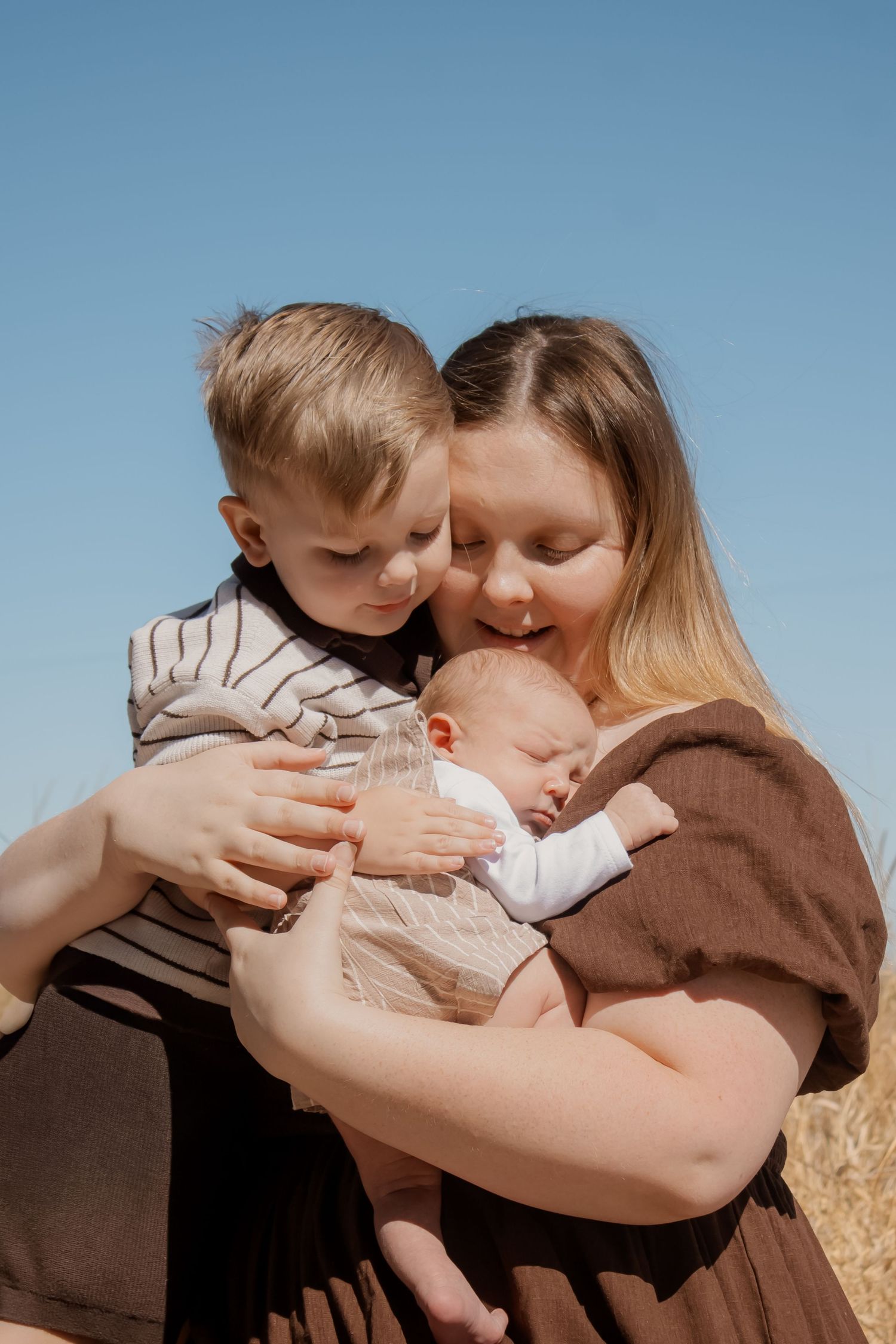 A mother holds a newborn baby and young boy outdoors under a clear blue sky in a warm family portrait.