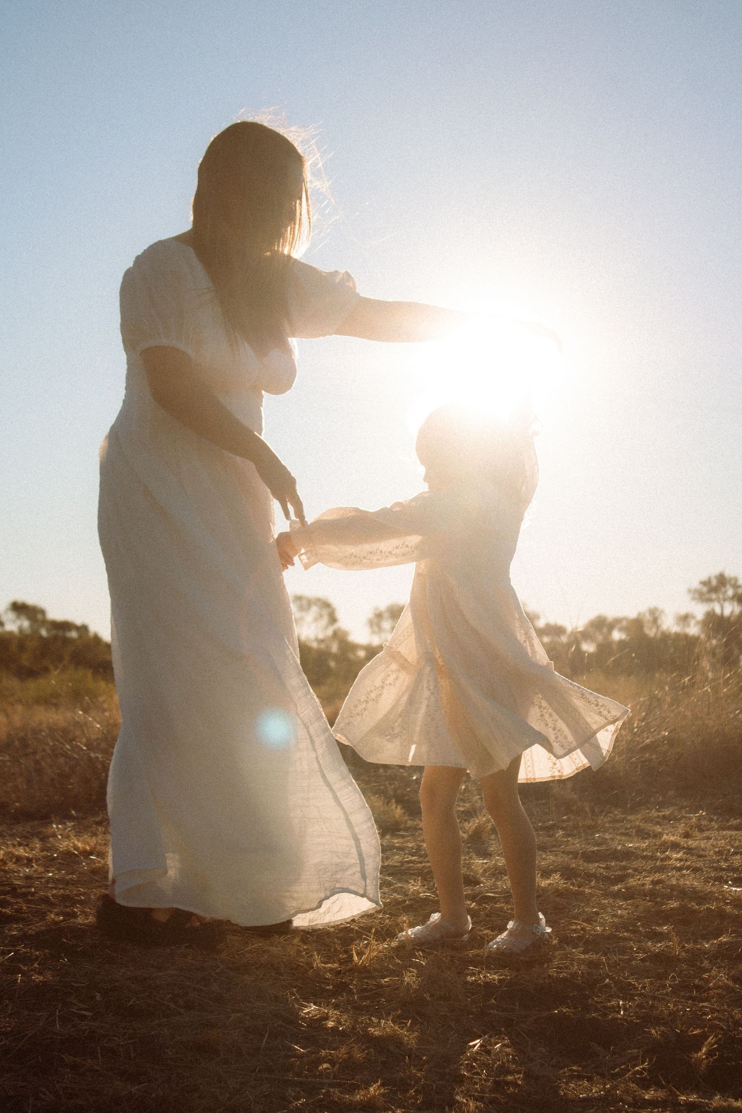 Mother and daughter in white dresses dancing in a sunlit field at golden hour.