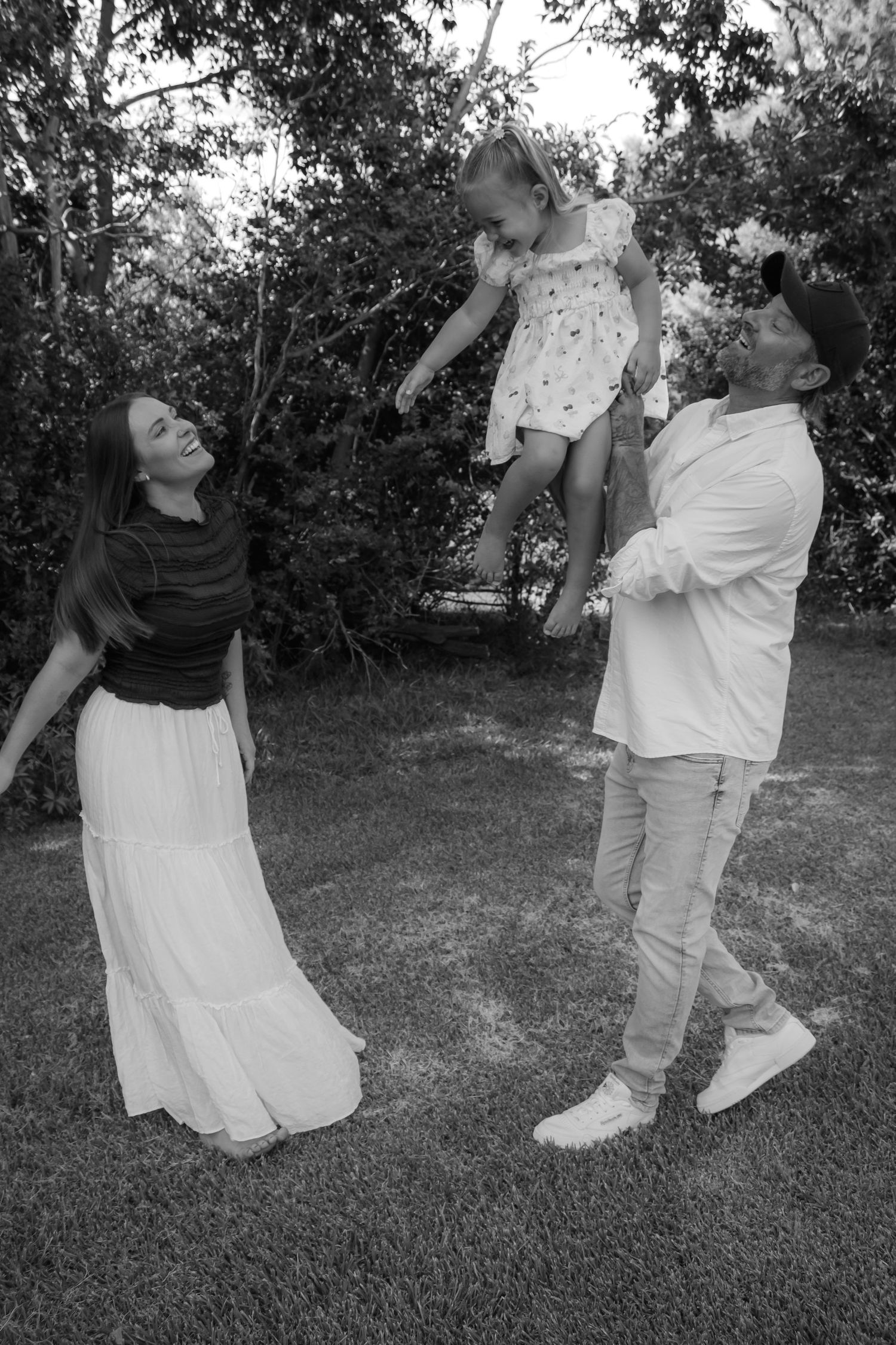 Black and white photo of a joyful family outdoors, with a man lifting a young girl in the air while a woman laughs beside them.