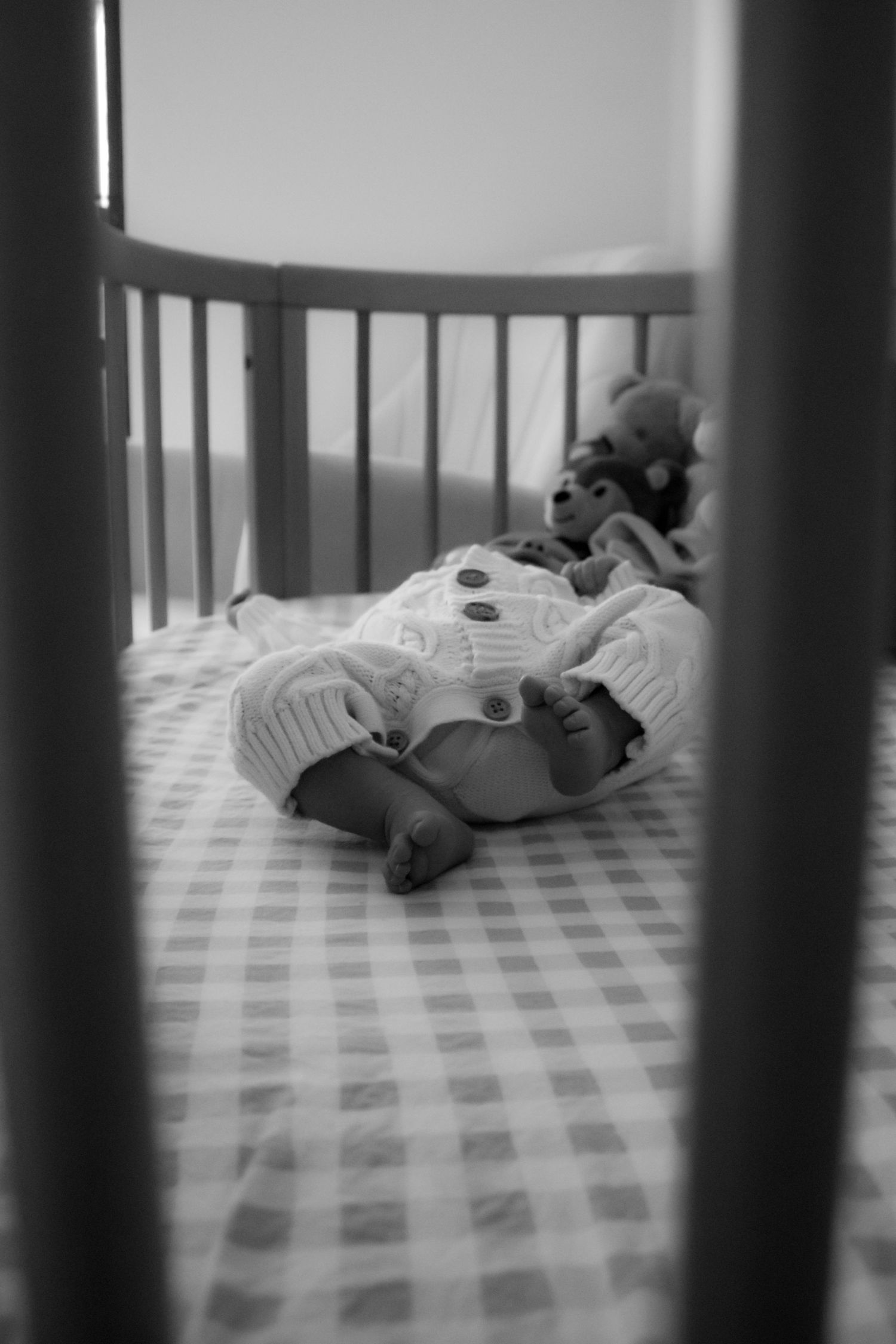 Black and white photo of a sleeping newborn baby in a crib with plaid sheets and a stuffed animal toy.