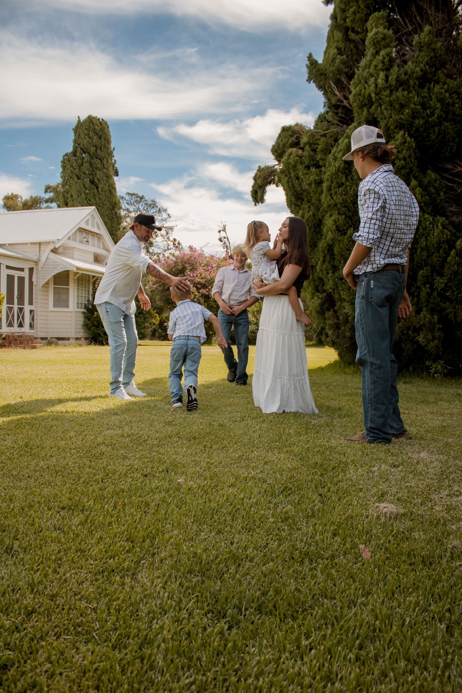 Family gathering outdoors on green lawn near white farmhouse, adults and children playing together under blue sky.