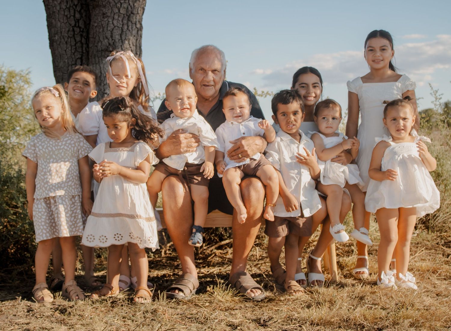 Large family group with grandfather and many grandchildren posing together outdoors under a tree in summer.