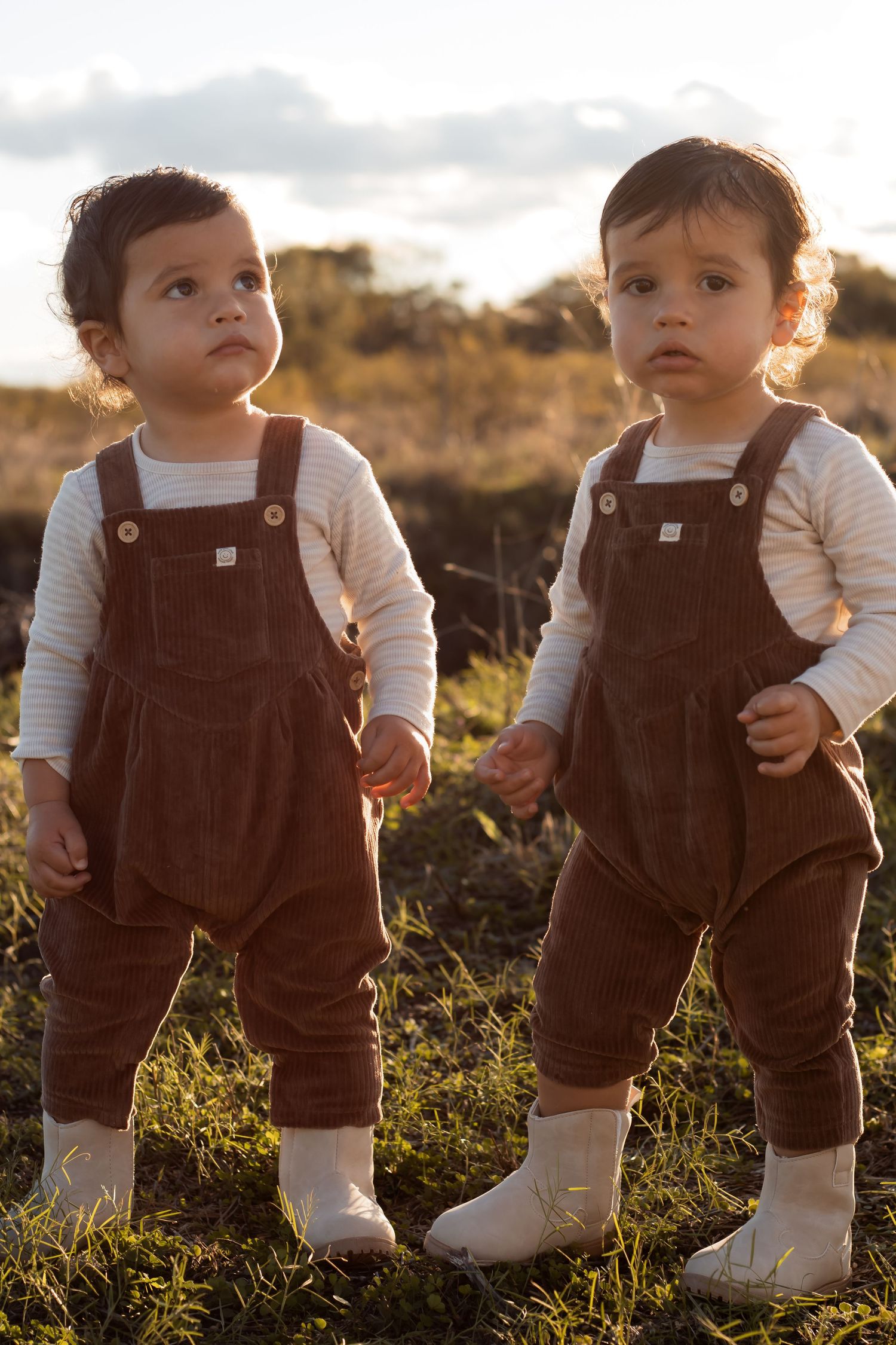 Twin toddlers in matching brown corduroy overalls standing together in a sunlit field.