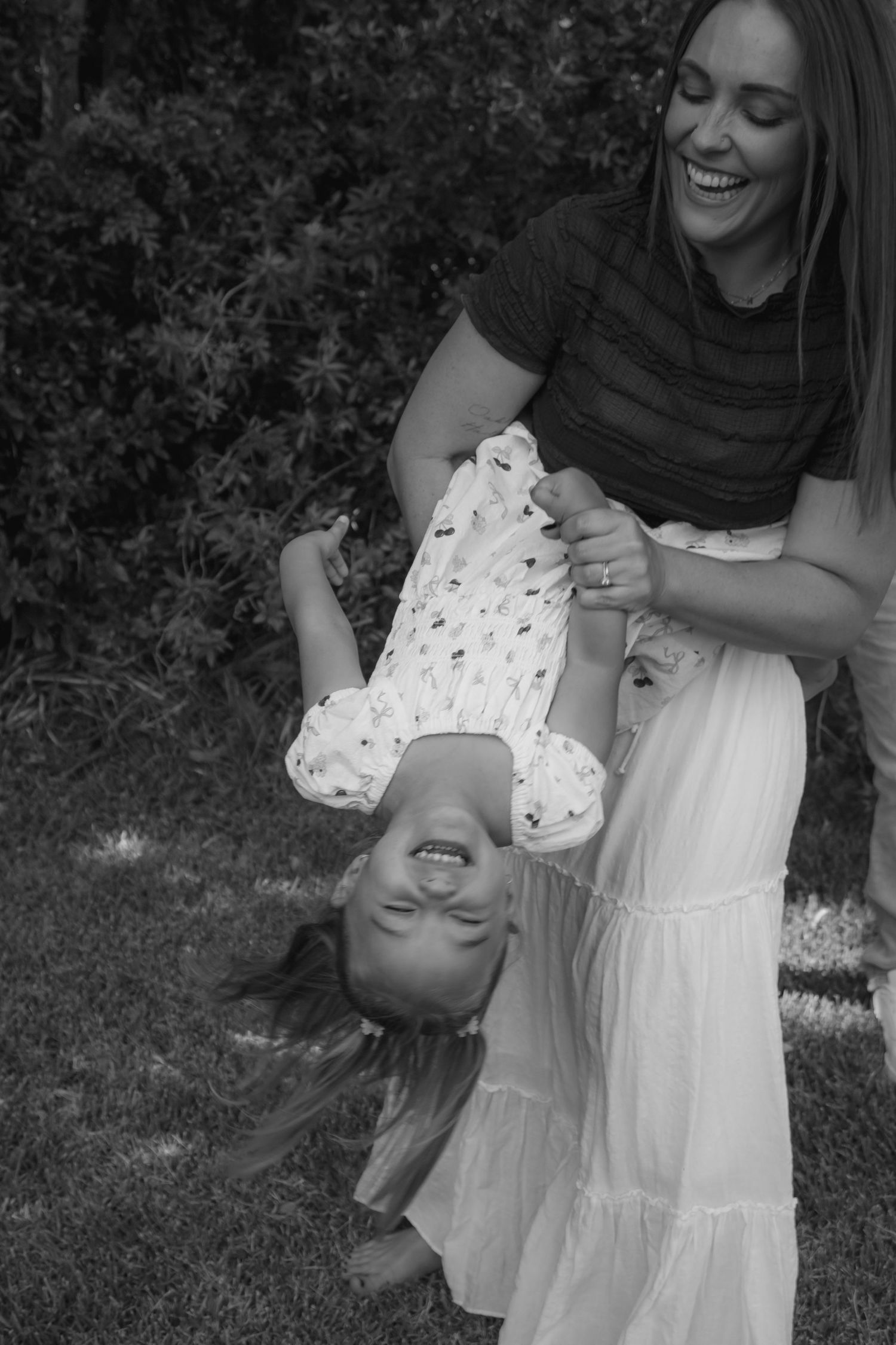 Black and white photo of a laughing mother playfully holding her young daughter upside down outdoors.
