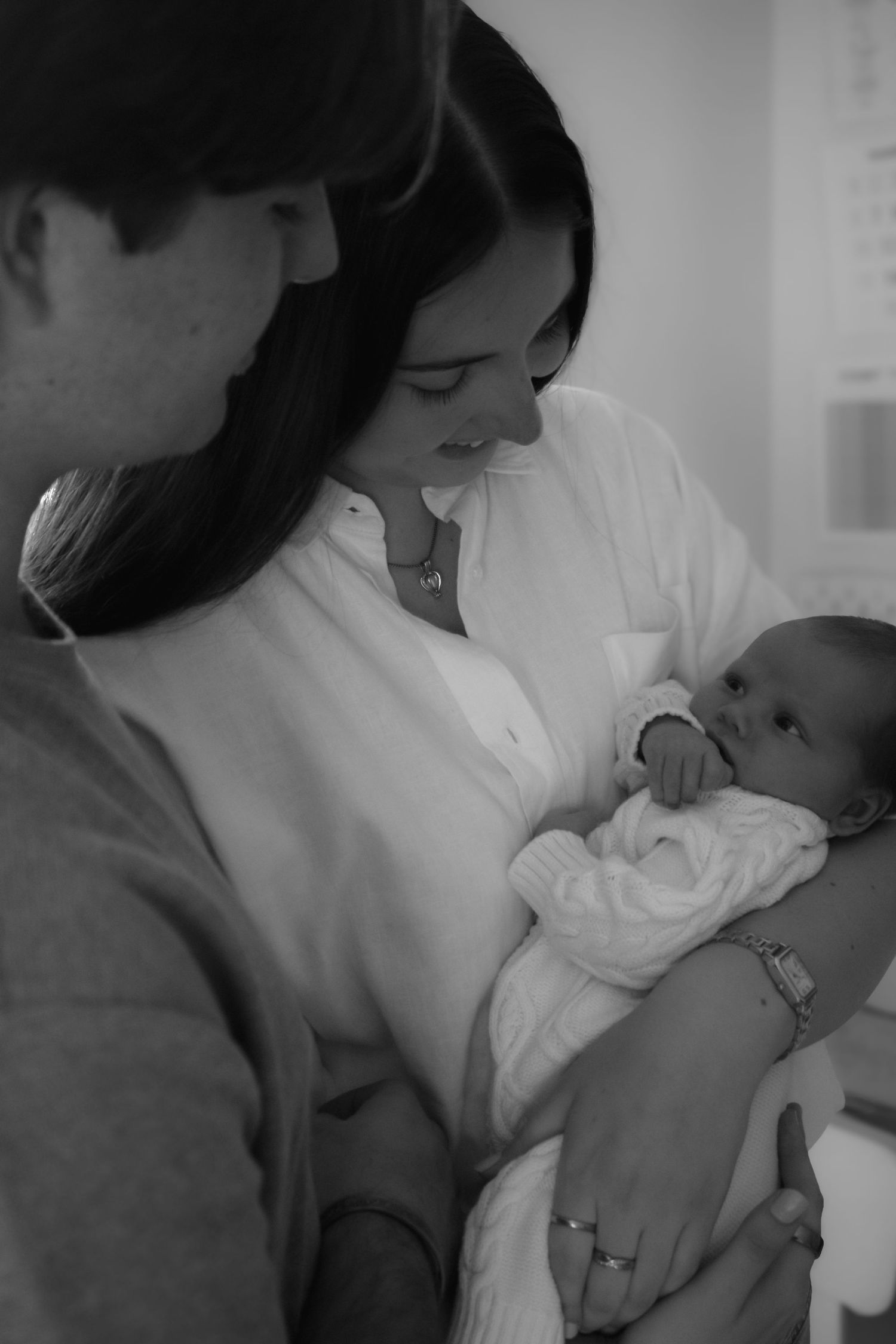 Black and white photo of new parents lovingly gazing at their newborn baby wrapped in a white blanket.