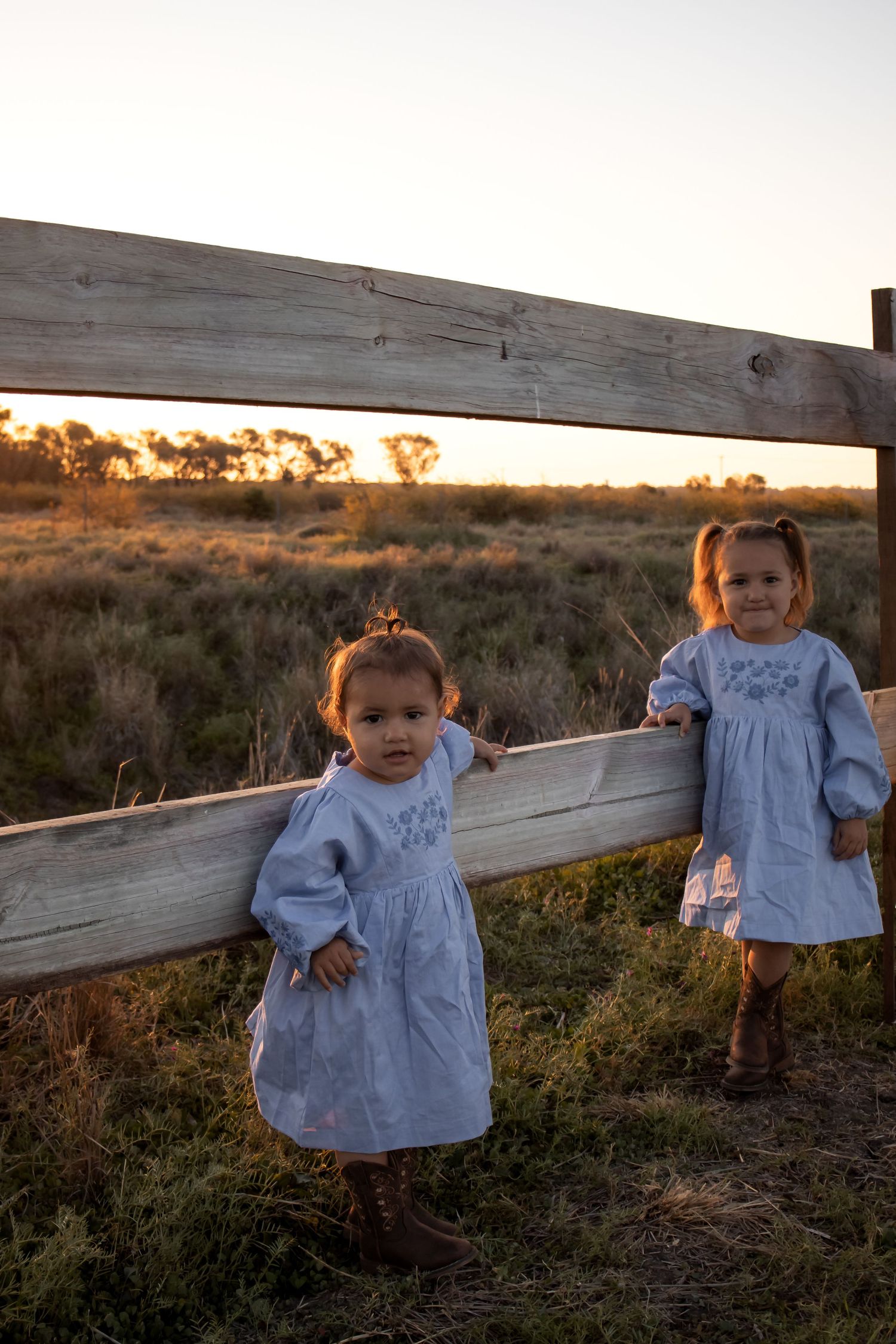 Two young girls in matching white dresses and boots stand by a wooden fence at sunset in a rural field.