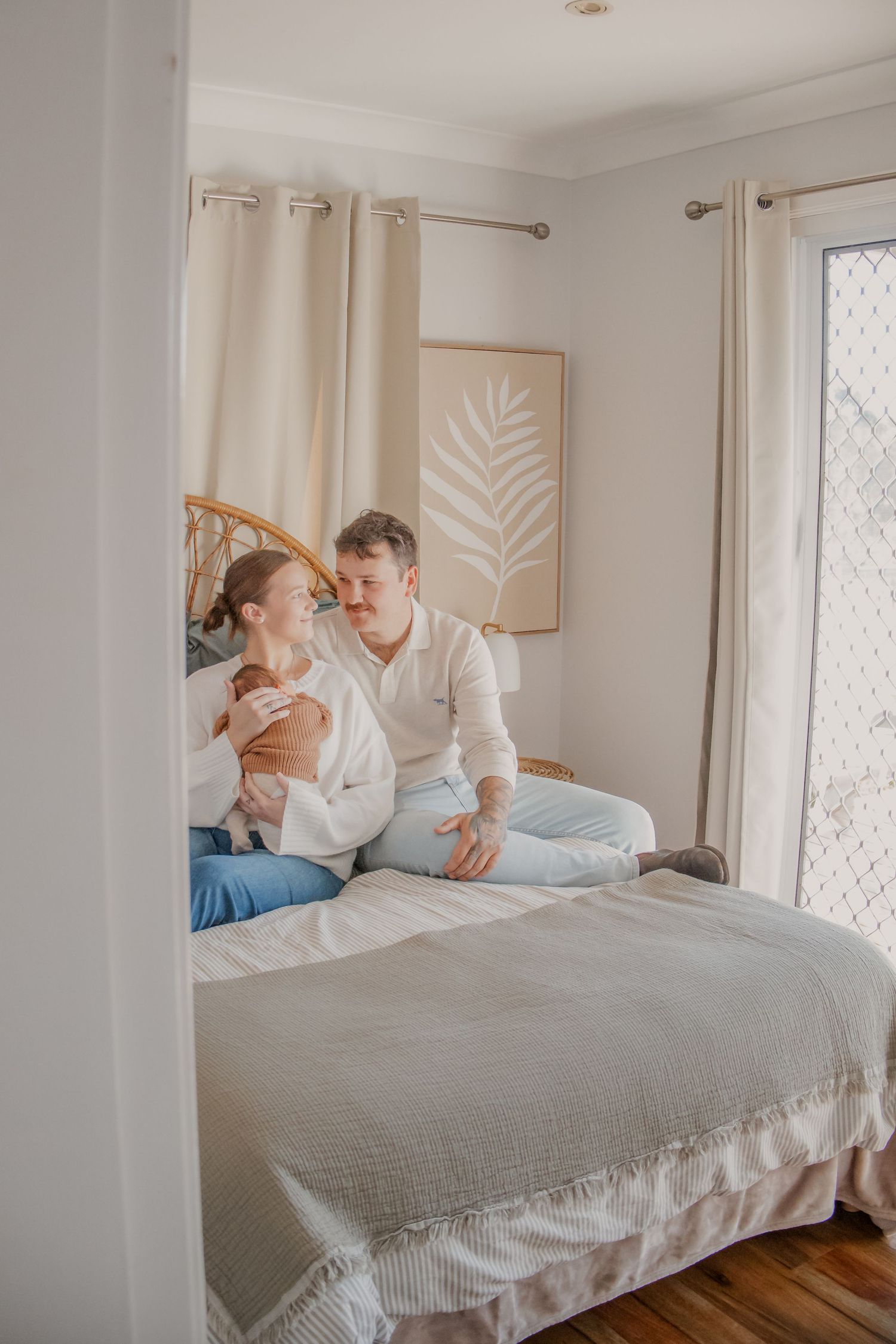 A couple sits on a bed holding their newborn baby in a bright, cozy bedroom with neutral decor.