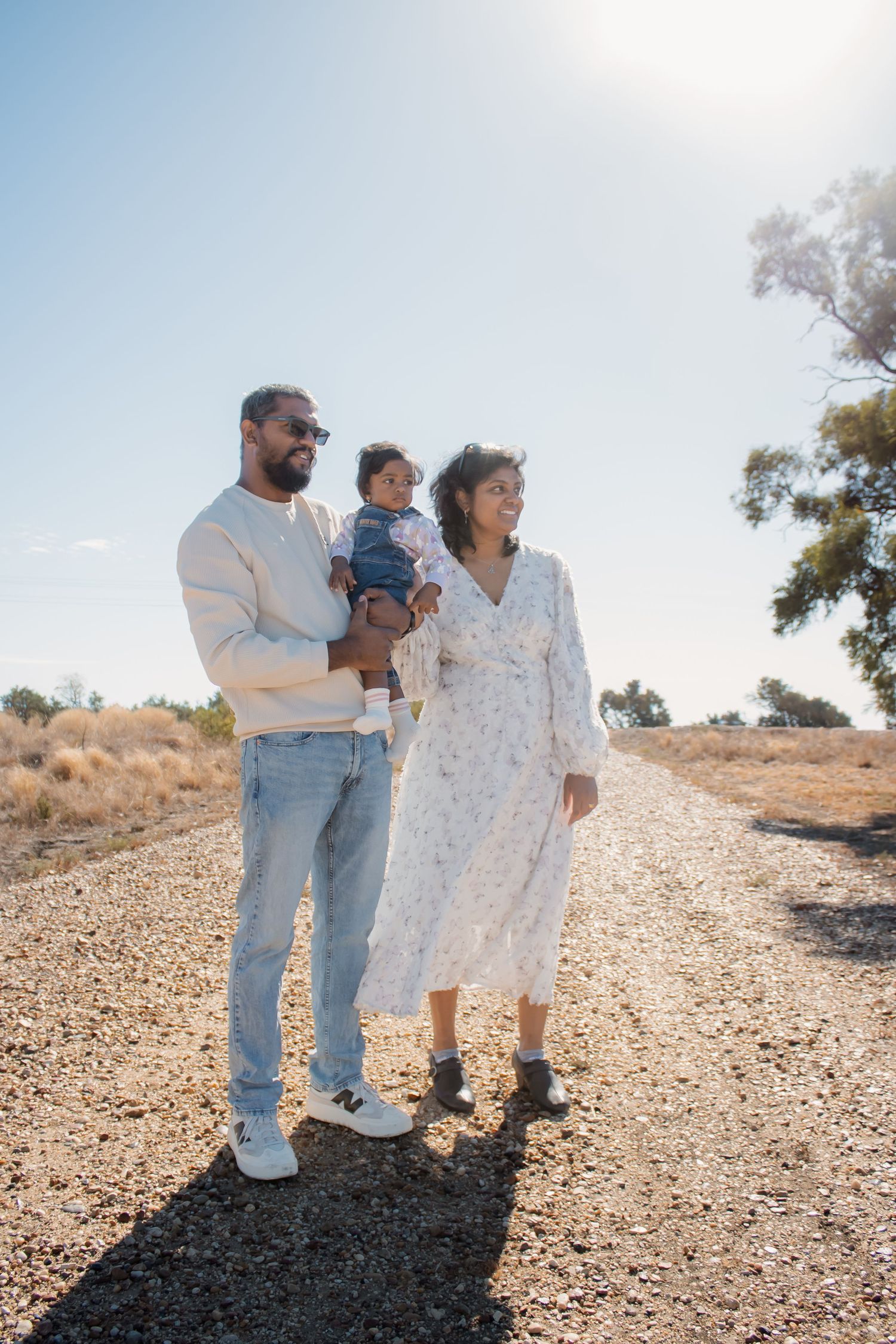 A family of three walks on a sunny gravel path, father holding baby, mother in floral dress, with blue sky behind.