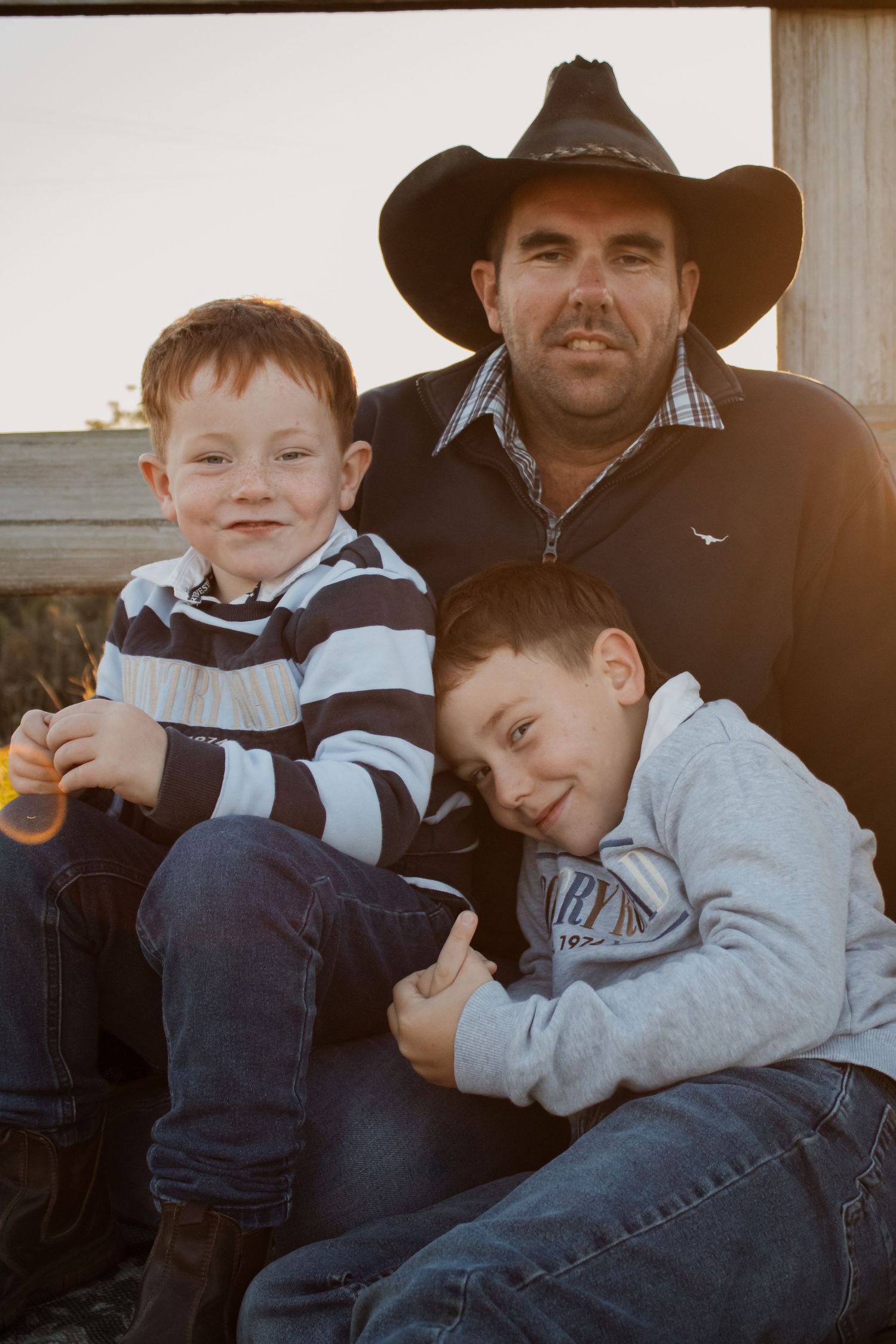 A man in a cowboy hat poses outdoors with two young boys at golden hour near a wooden fence.