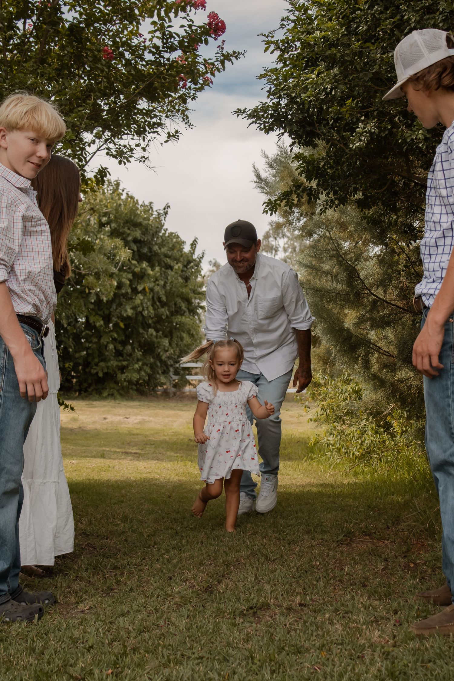 Family gathering outdoors with adults and children playing on green grass surrounded by lush trees.