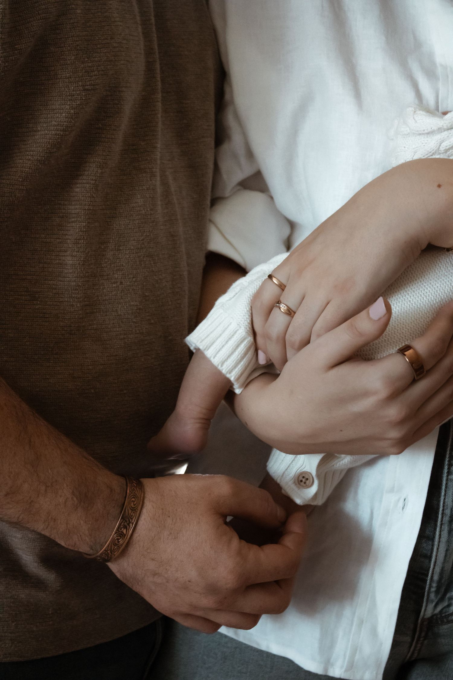 Close-up of a couple holding hands, wearing gold rings and a leather bracelet, dressed in white and brown tops.