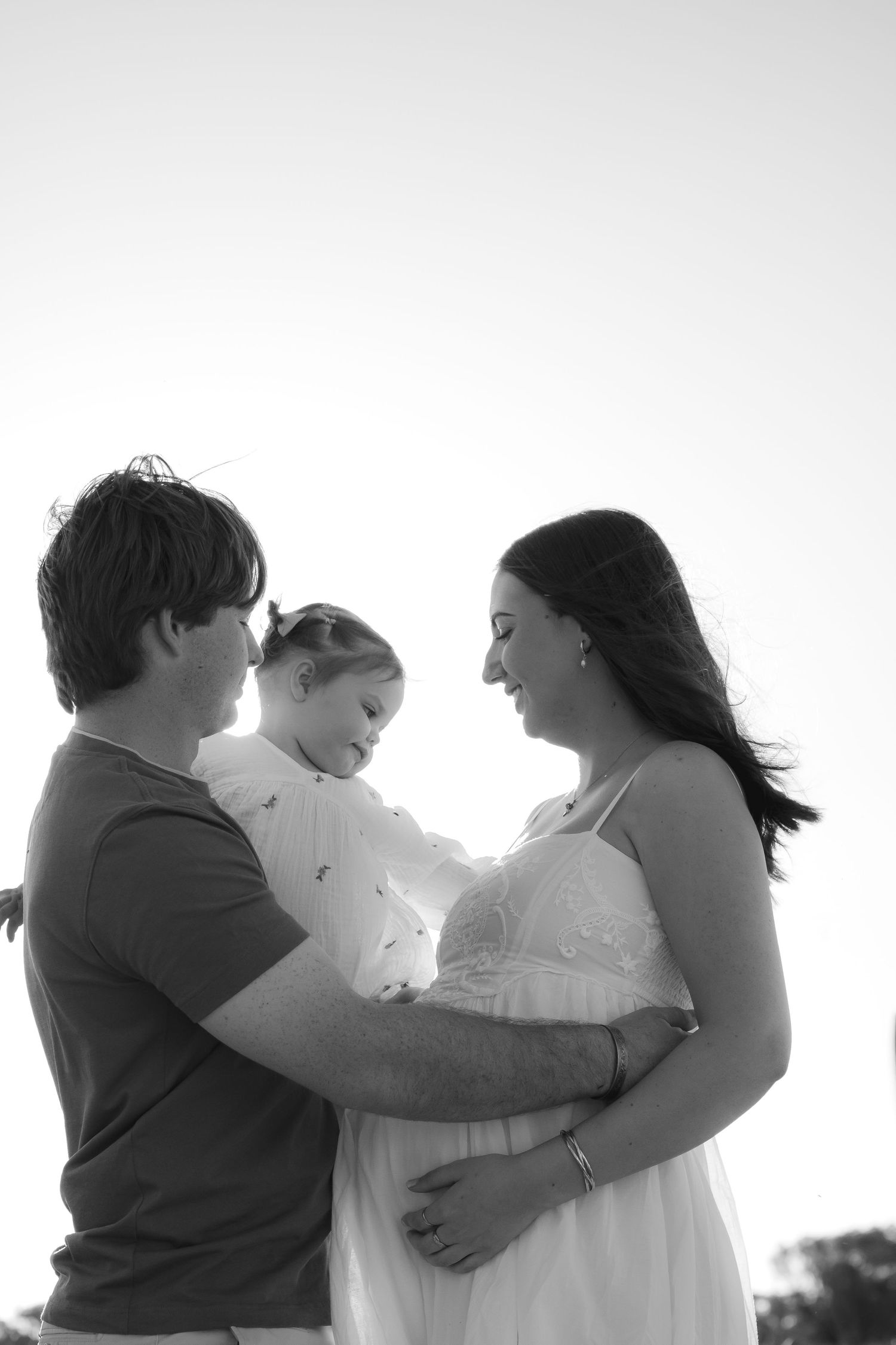 Black and white photo of a couple holding their young child outdoors, with the pregnant mother in a white dress.