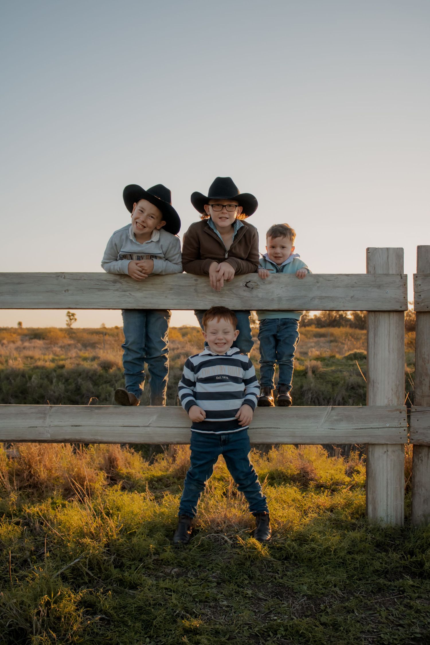 Four young boys in cowboy hats pose on a wooden ranch fence at golden hour in an open field.