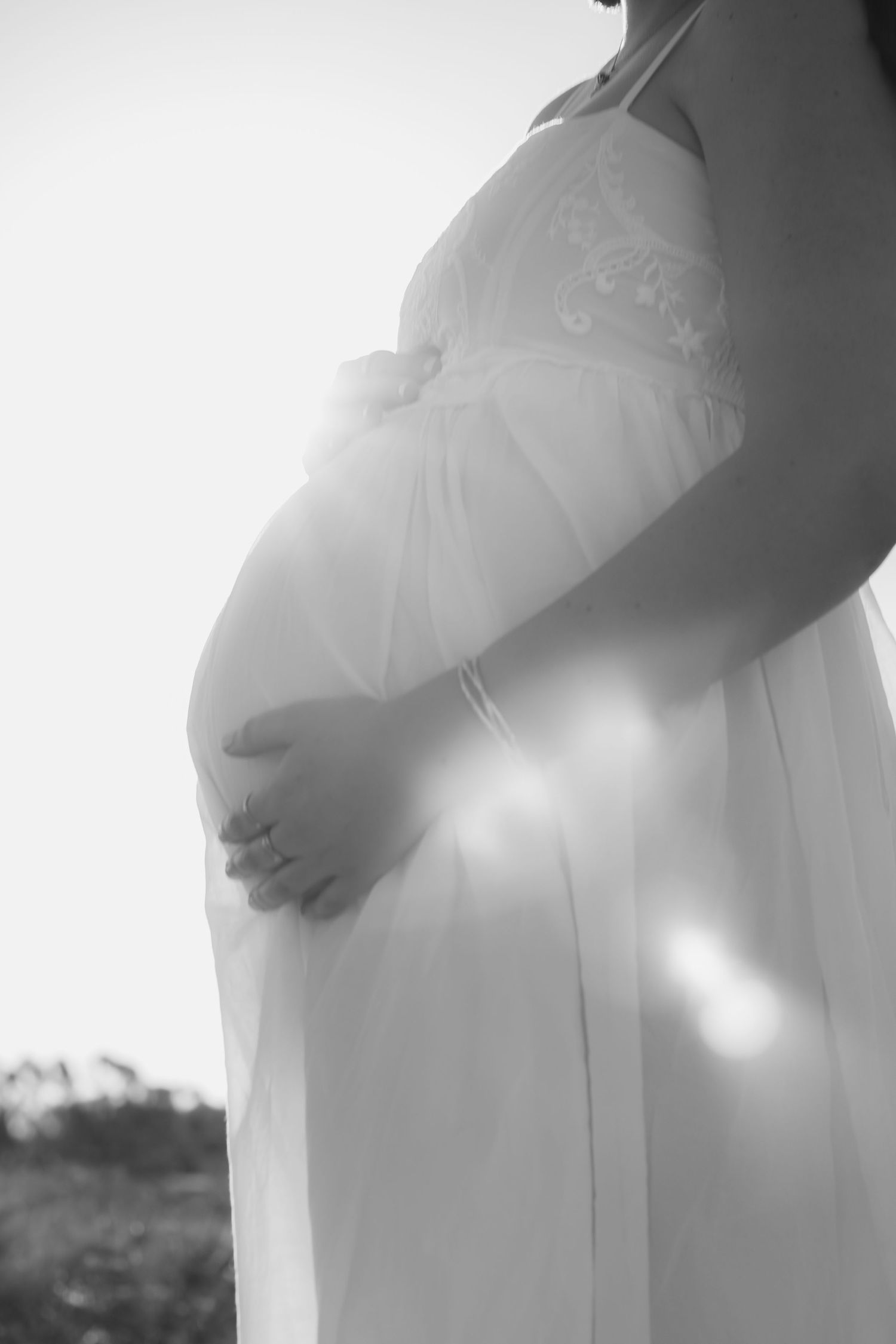 Black and white close-up of a pregnant woman in a flowing white dress cradling her baby bump outdoors.