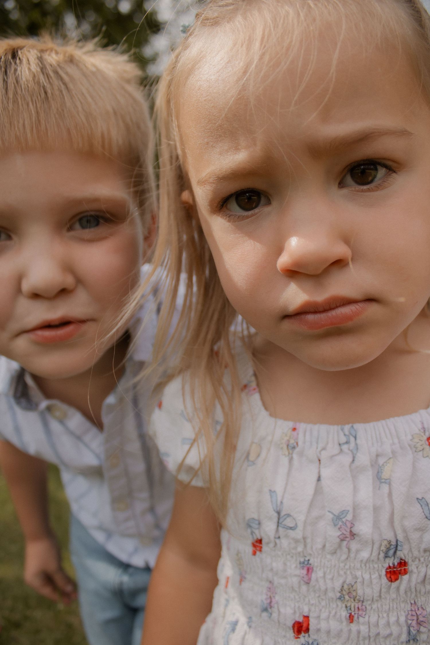 Two young blonde children with sad expressions looking at camera outdoors.