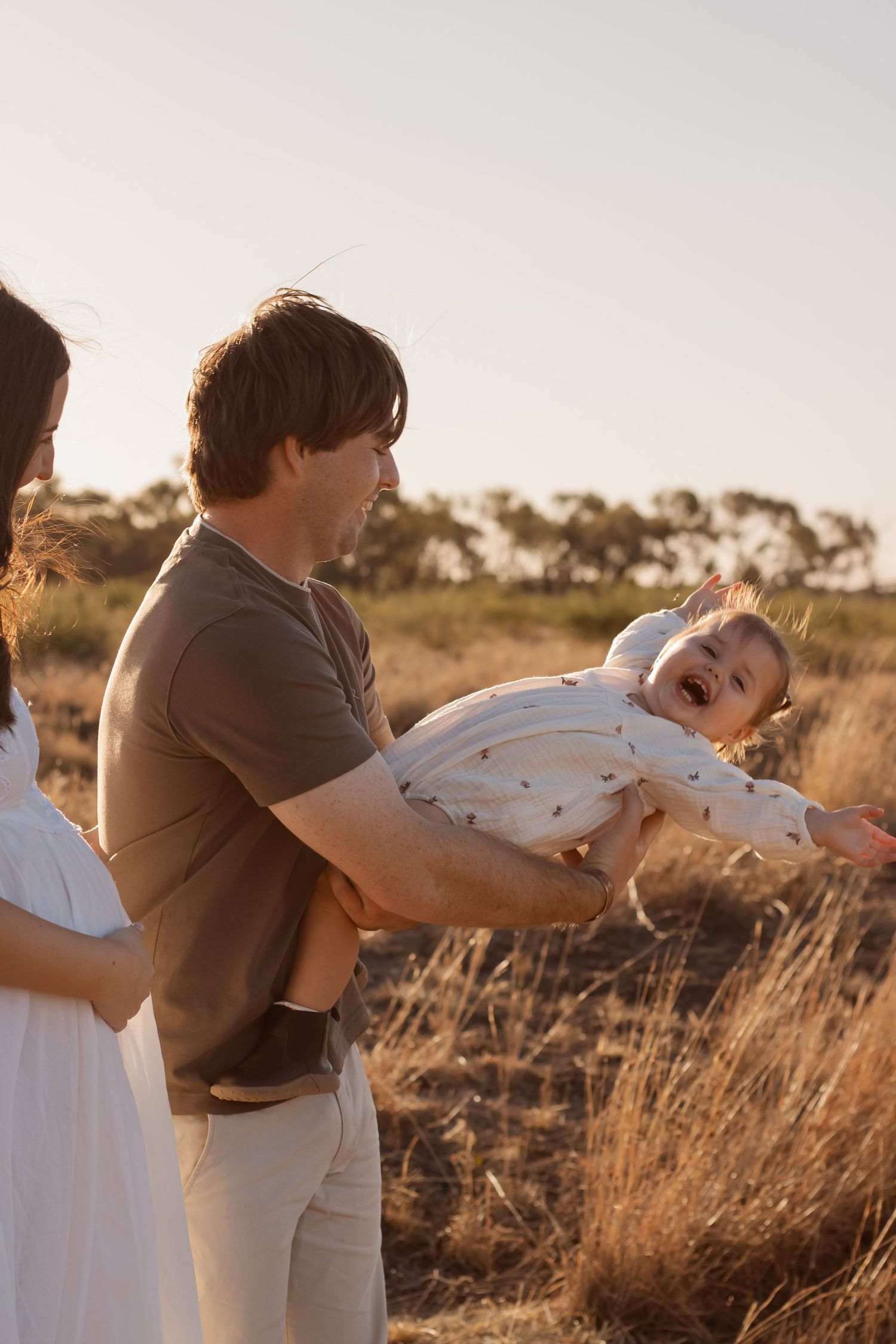 Father holding giggling baby outdoors in golden hour light with dry grass field backdrop.