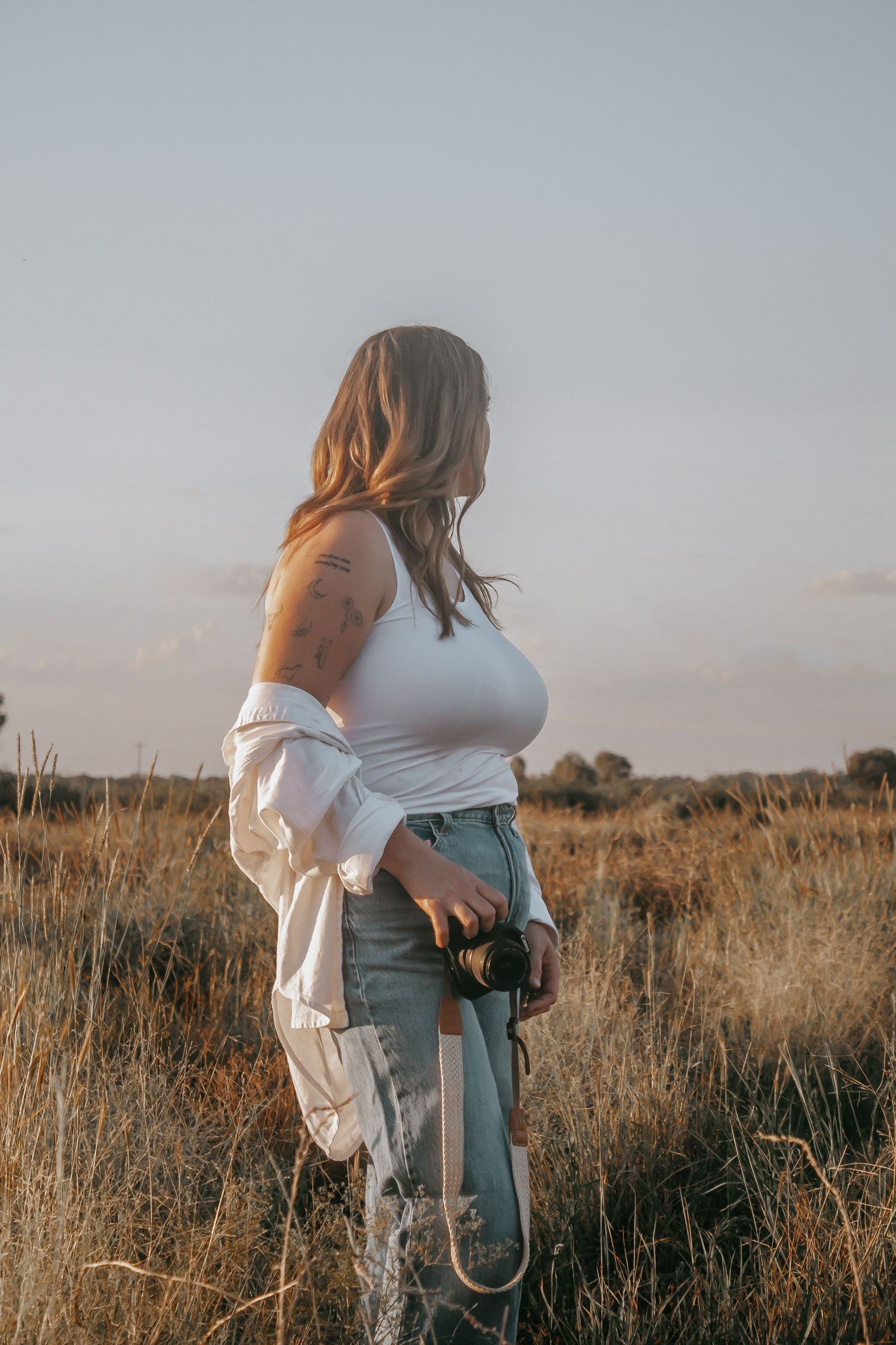 Woman in white top and jeans holding a camera standing in a golden field at sunset.