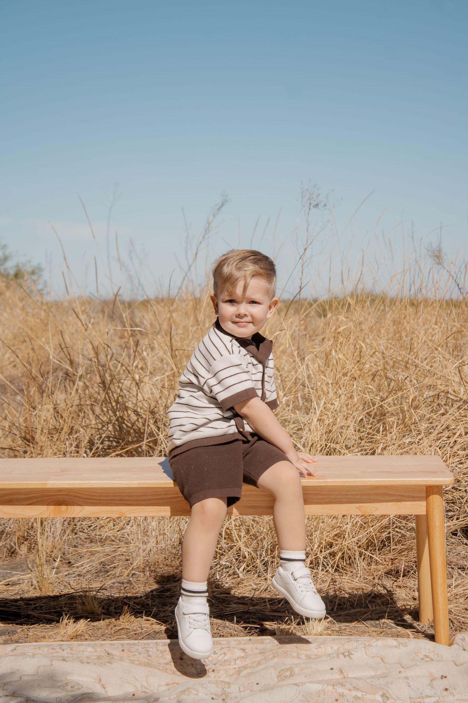 Young blond boy in striped polo and brown shorts sits on wooden bench in dry grassy field under blue sky.