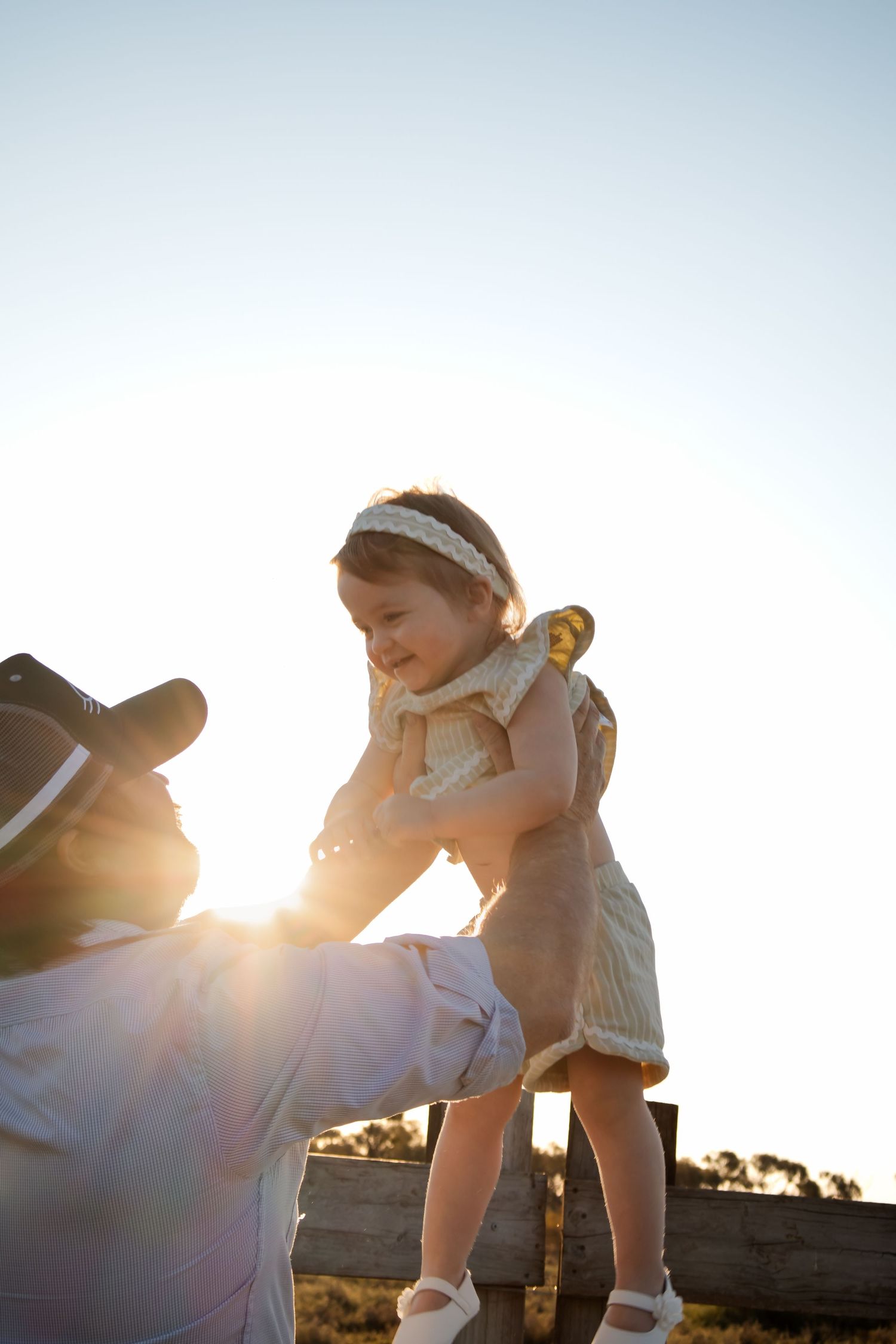 Adult lifting a smiling toddler girl over a wooden fence in golden sunset light outdoors.