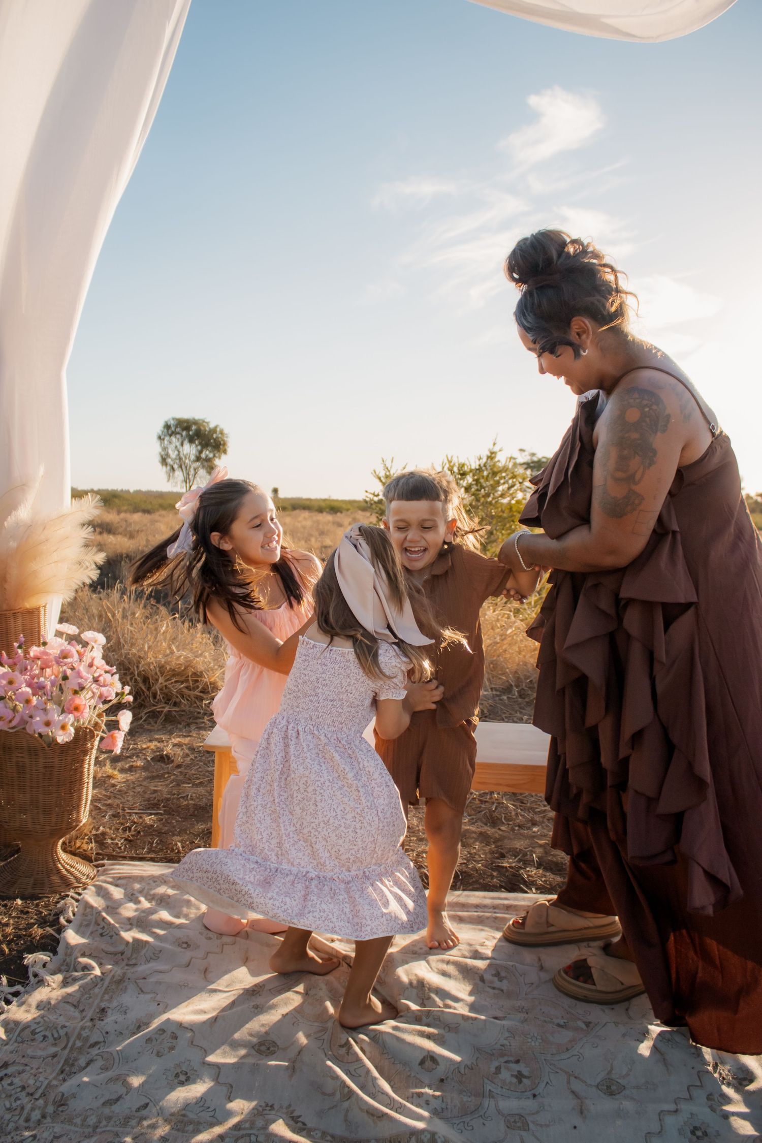 Three women dancing joyfully outdoors at a beach ceremony, wearing floral and brown dresses in golden sunlight.