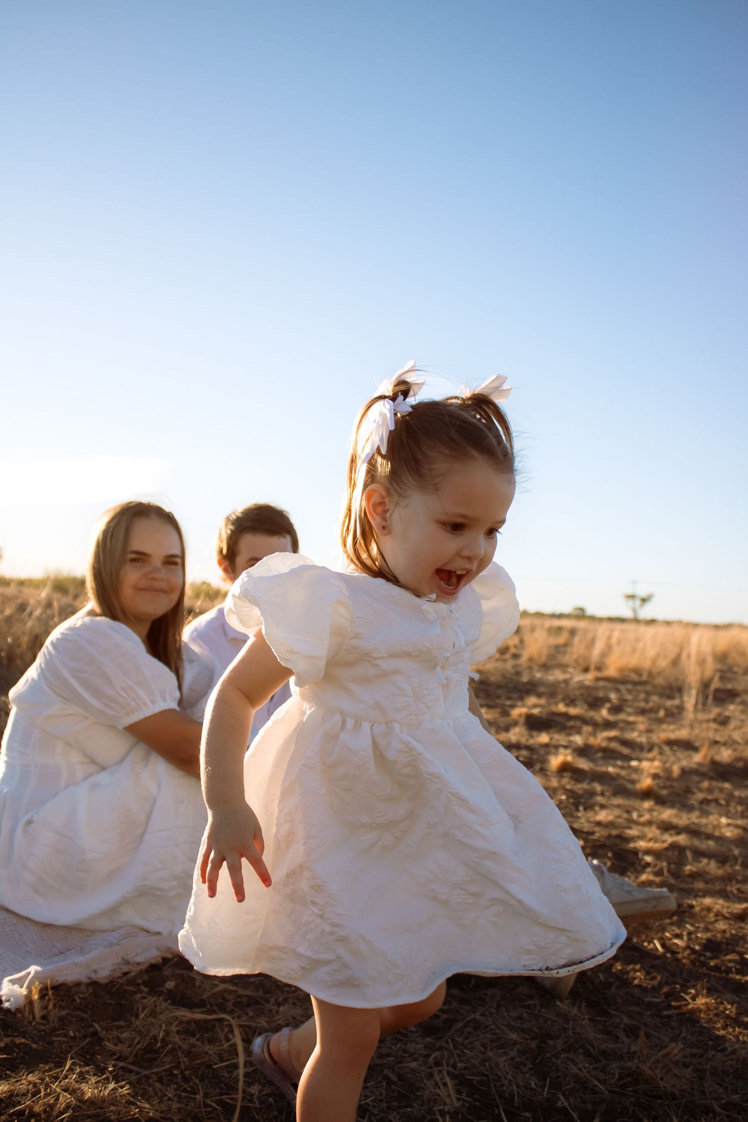 Young girl in white dress playing in golden field with two children in white outfits under clear blue sky.