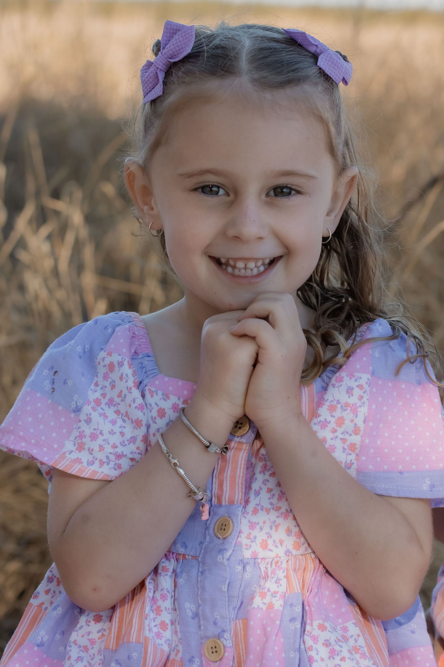 Smiling young girl in a pastel patchwork dress with purple hair bows, clasping hands near chin in a golden field.