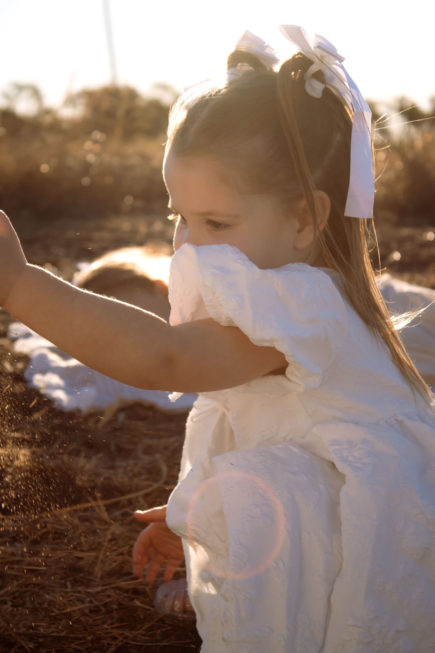 Young girl in white dress with bow playing outdoors in warm golden sunset light.