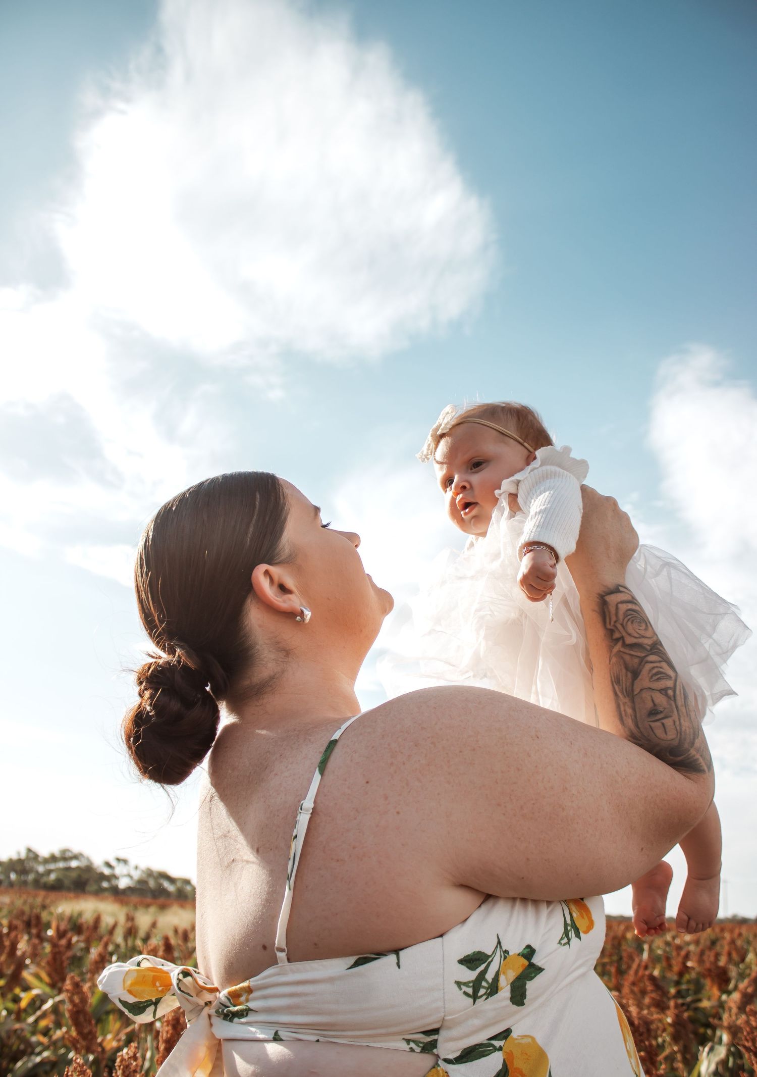 A tattooed mother in a floral dress lifts her baby girl outdoors under a bright blue sky.