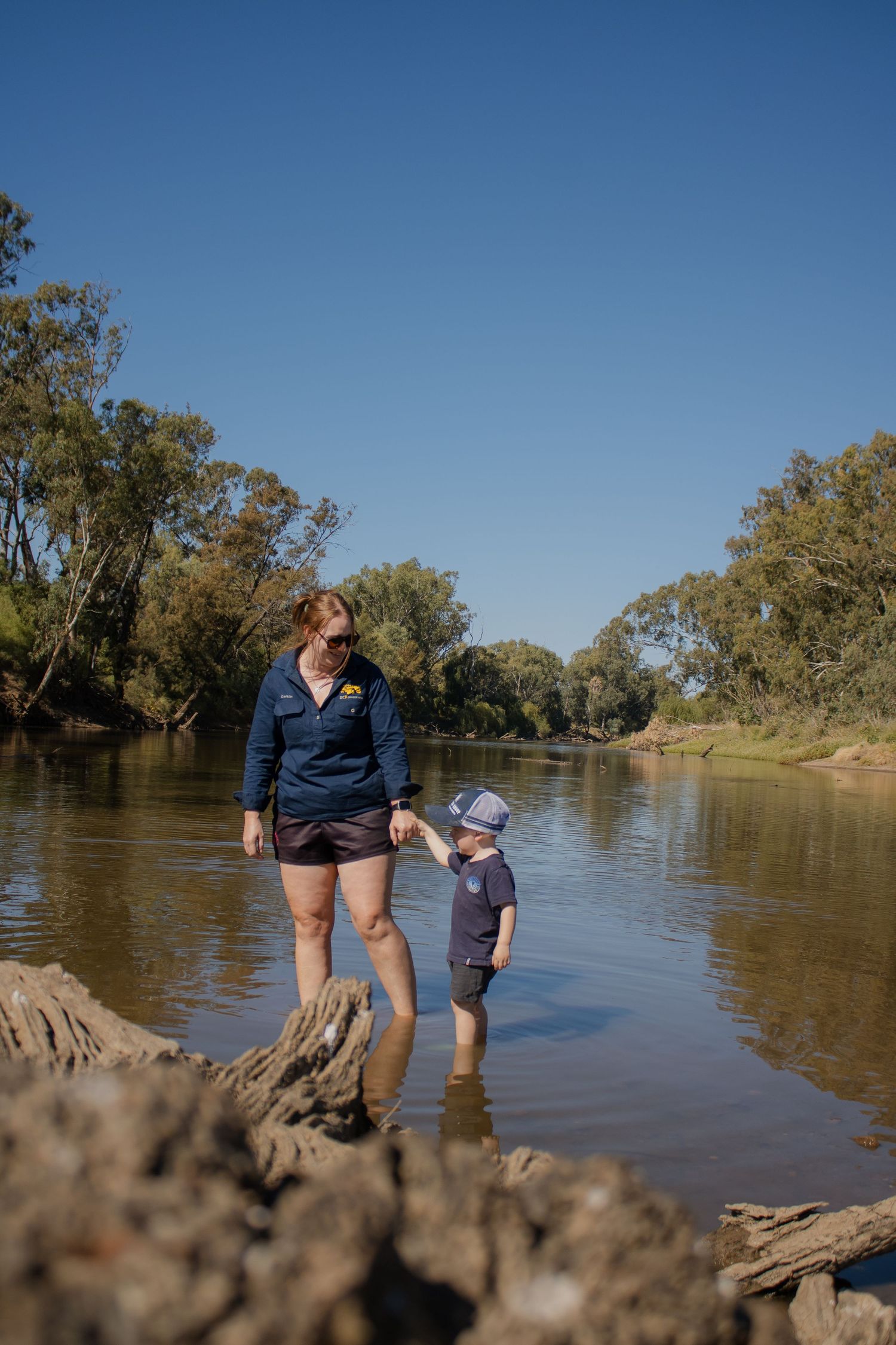 A woman and toddler wade in a shallow Australian river surrounded by gum trees under a clear blue sky.
