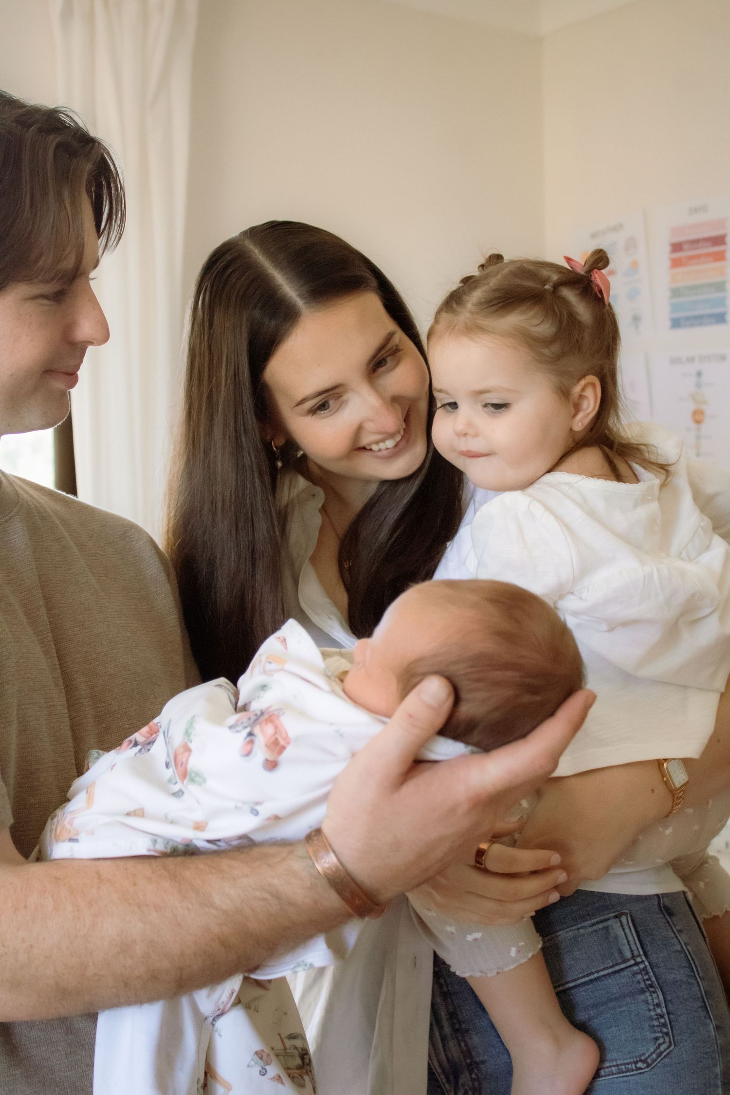 Happy family with parents and toddler girl admiring their newborn baby wrapped in a swaddle blanket indoors.