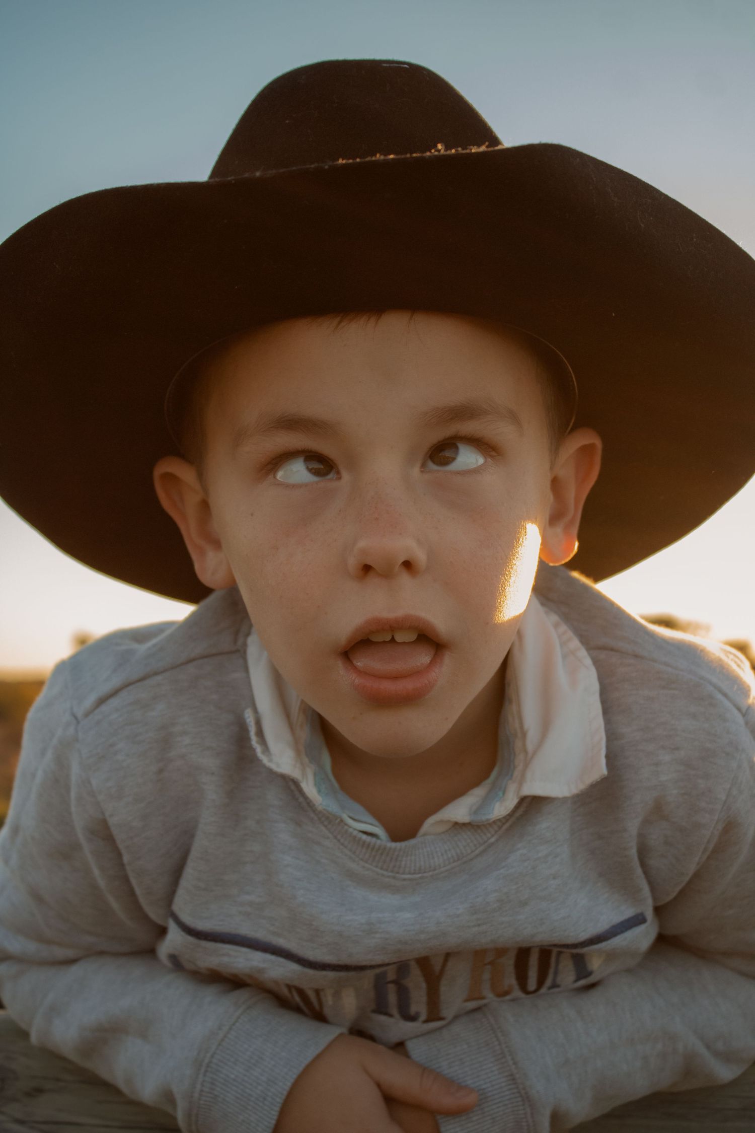 Young boy wearing a black cowboy hat outdoors at golden hour, looking up with a curious expression.