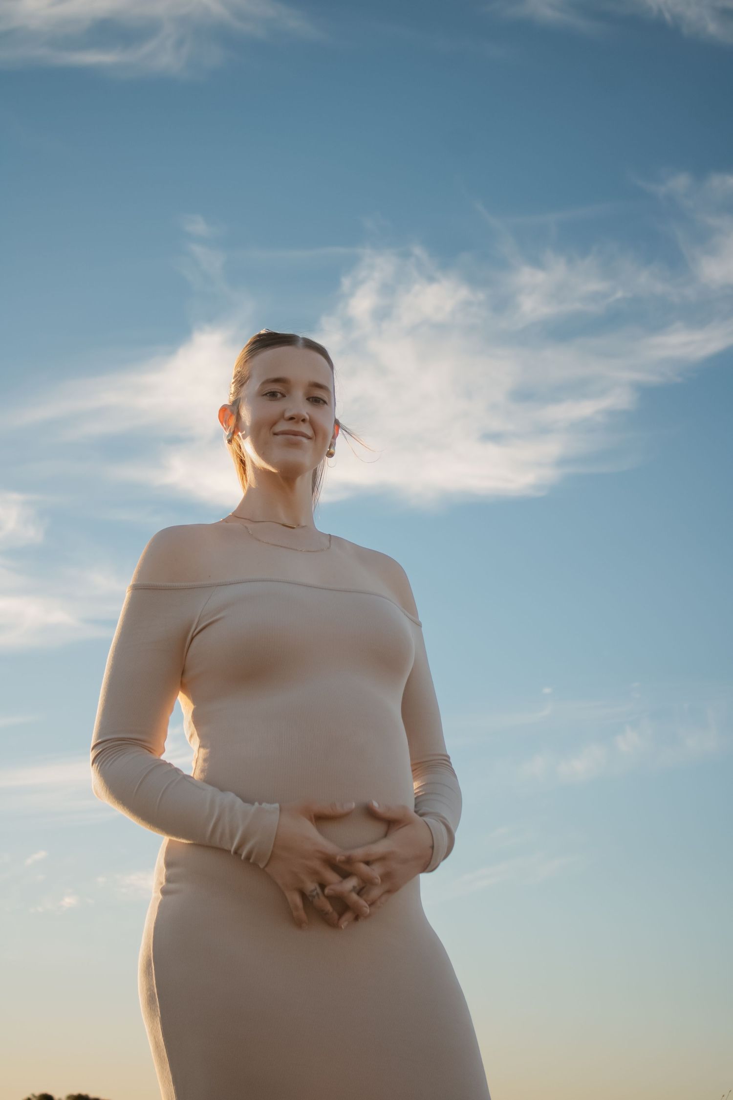 Pregnant woman in beige off-shoulder dress stands outdoors against blue sky, smiling with hands resting on belly.