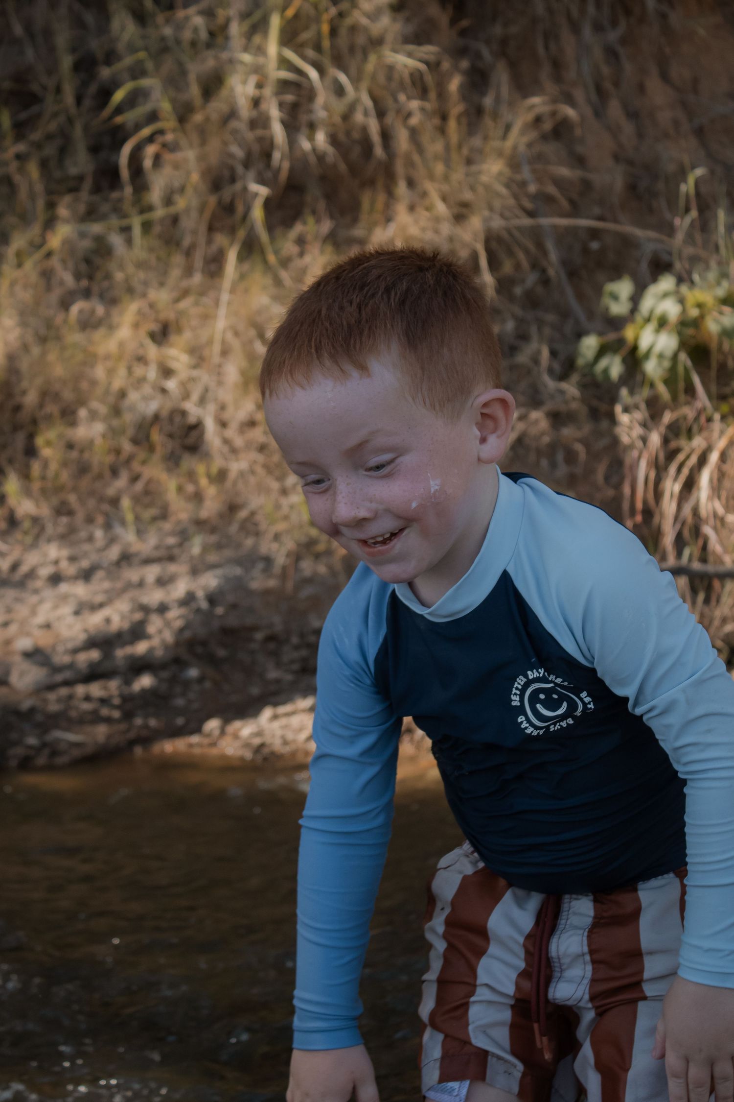 Smiling red-haired boy in rash guard shirt playing near a creek outdoors.
