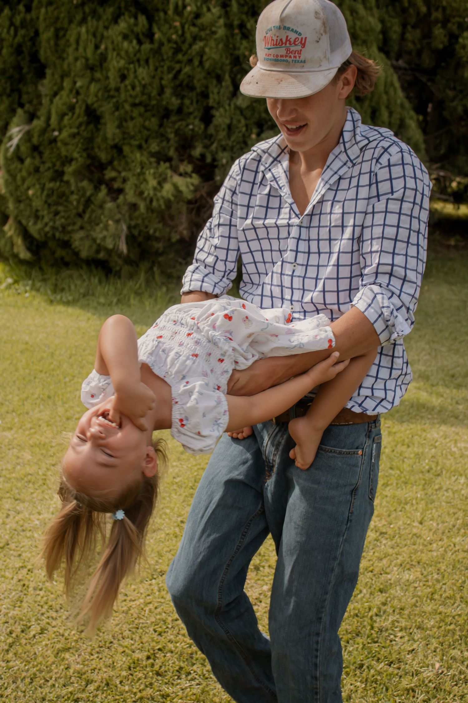 Parent playfully holding child upside down while laughing outdoors on green grass near evergreen trees.