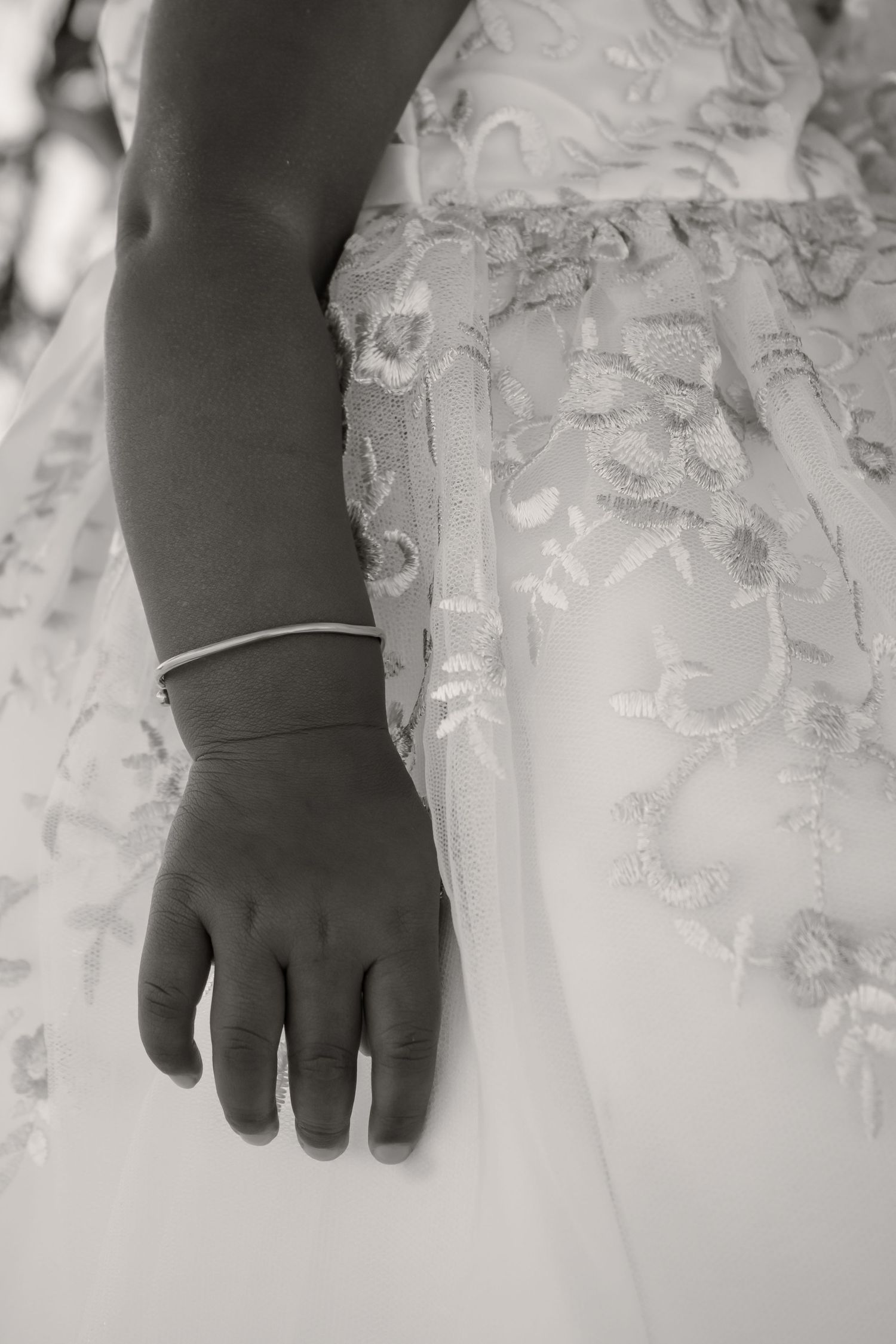 Black and white photo of a child's hand with a delicate bracelet resting against an embroidered white lace wedding dress.
